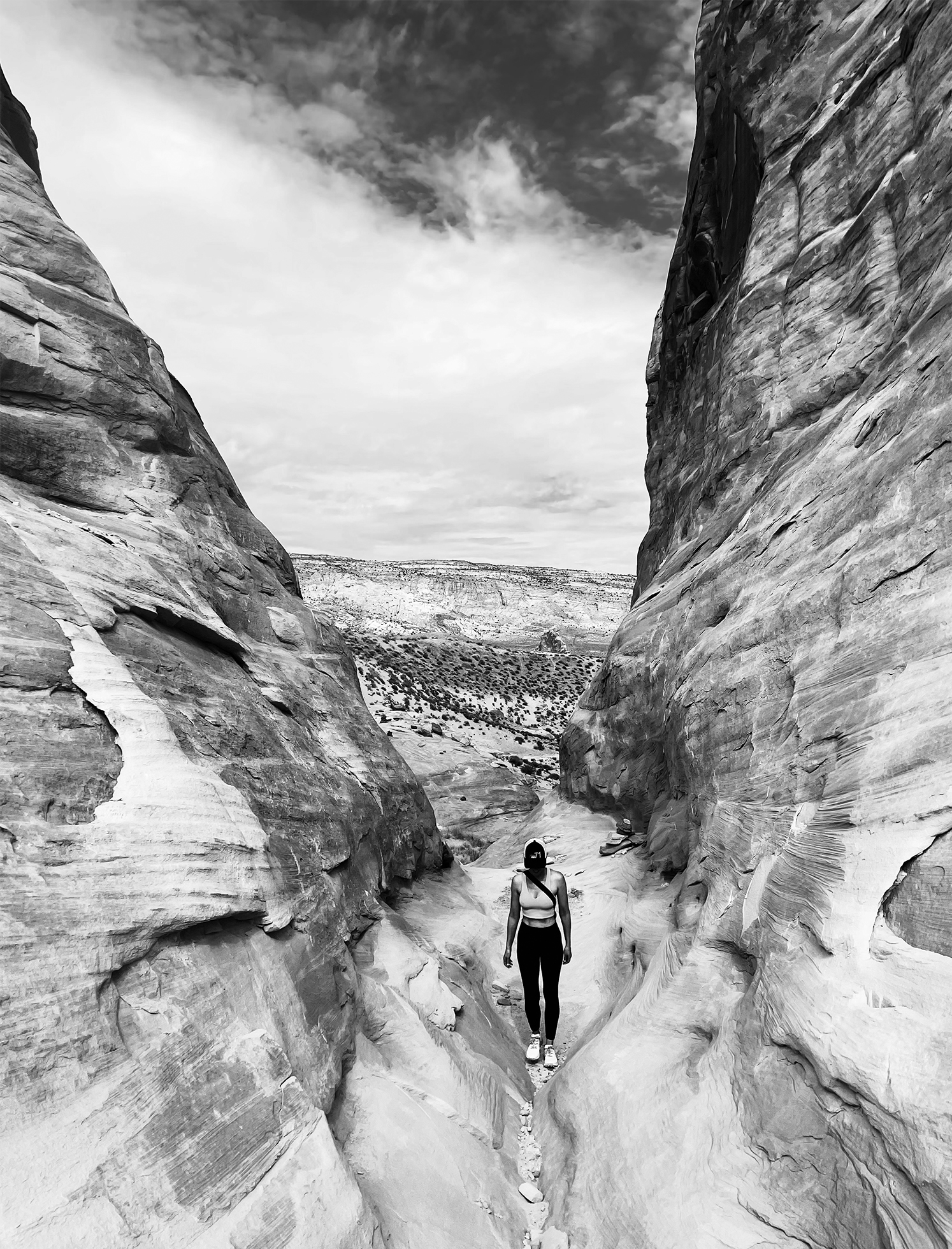 A fit woman in black-and-white trendy sports gear looks down as she walks toward the camera and along the center of a narrow opening between two tall, vertical rock formations.