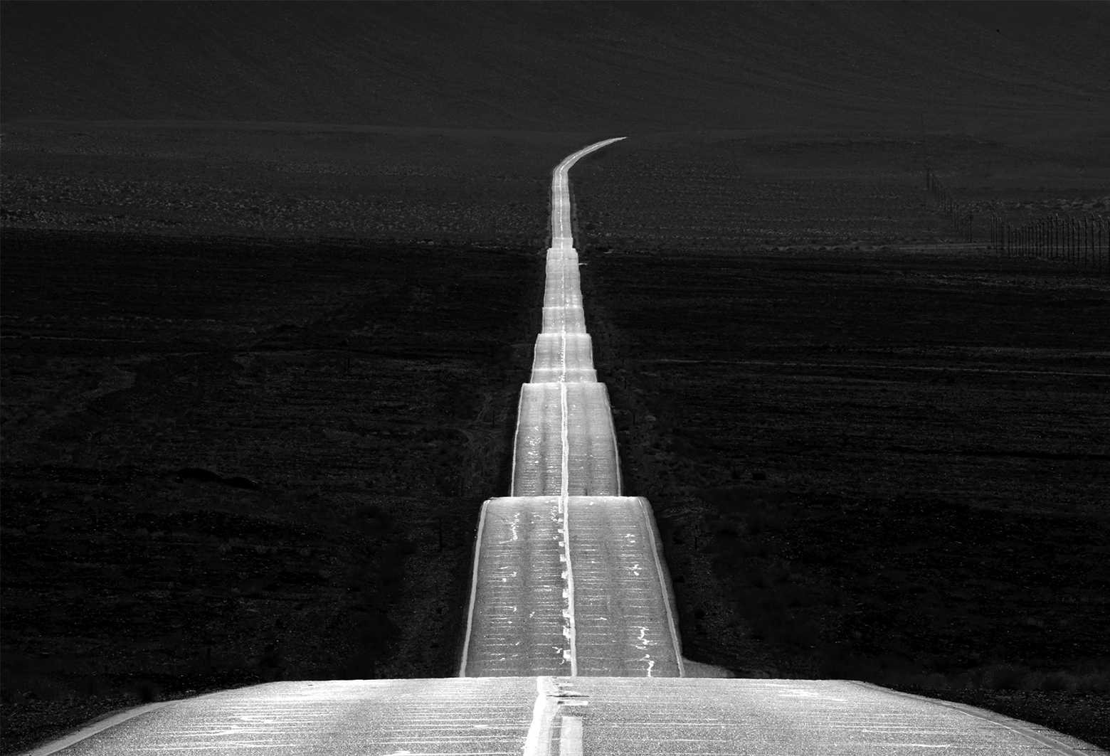 A two-lane road to nowhere with its highest point in the foreground undulates ribbon-like down and across the center of a desolate landscape tapering off and stretching into the horizon.