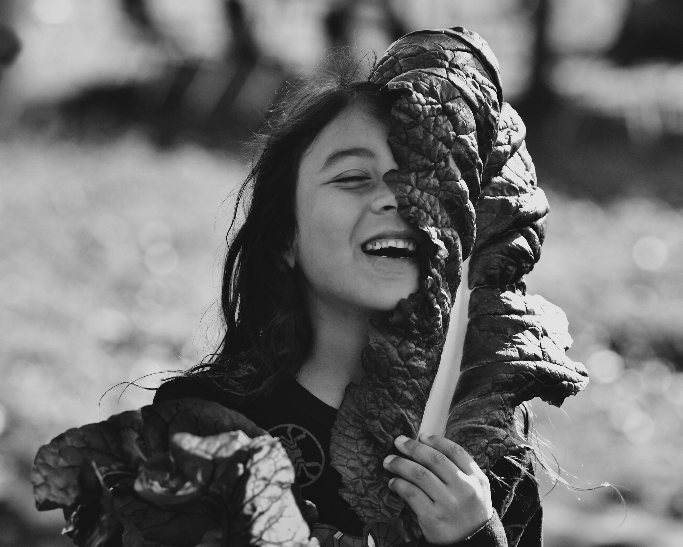 A young girl smiles joyously with her eyes closed as she almost caresses, as if it were a feather, a very large very long kale leaf that reaches all the way from her chest and curls up and over the top of her head hiding half of her face.