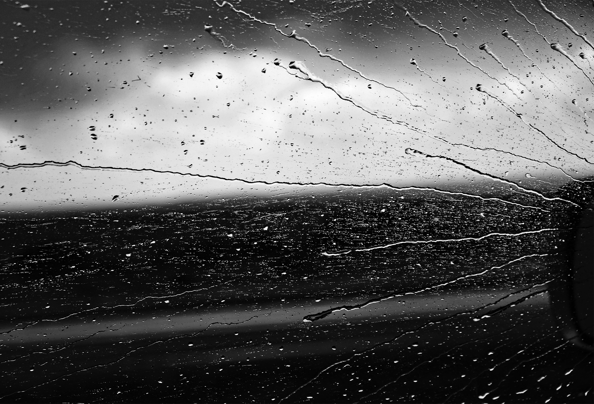 Rain streaks from right to left across a driver’s side car window viewed from inside a moving car. A small portion of the side mirror on the left is barely visible against the cloudy gray sky and the dark landscape.