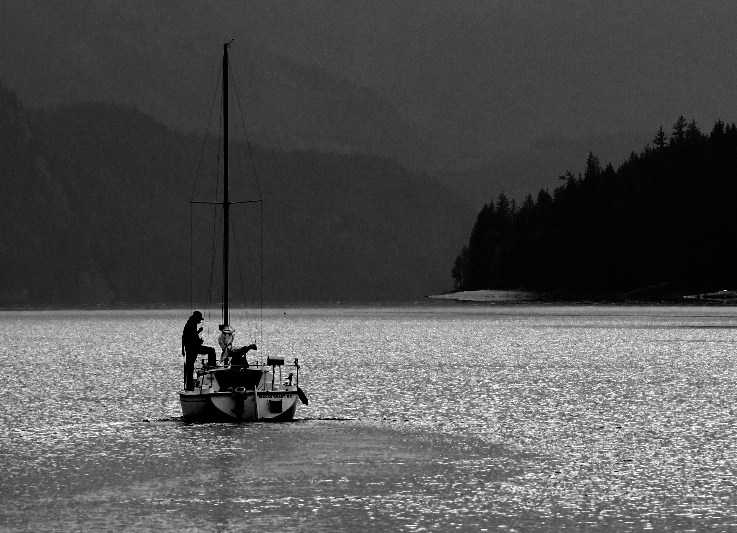 Sailboat at sunset with a person in silhouette on deck and the shoreline of Slocan Lake in British Columbia, Canada, in the background.