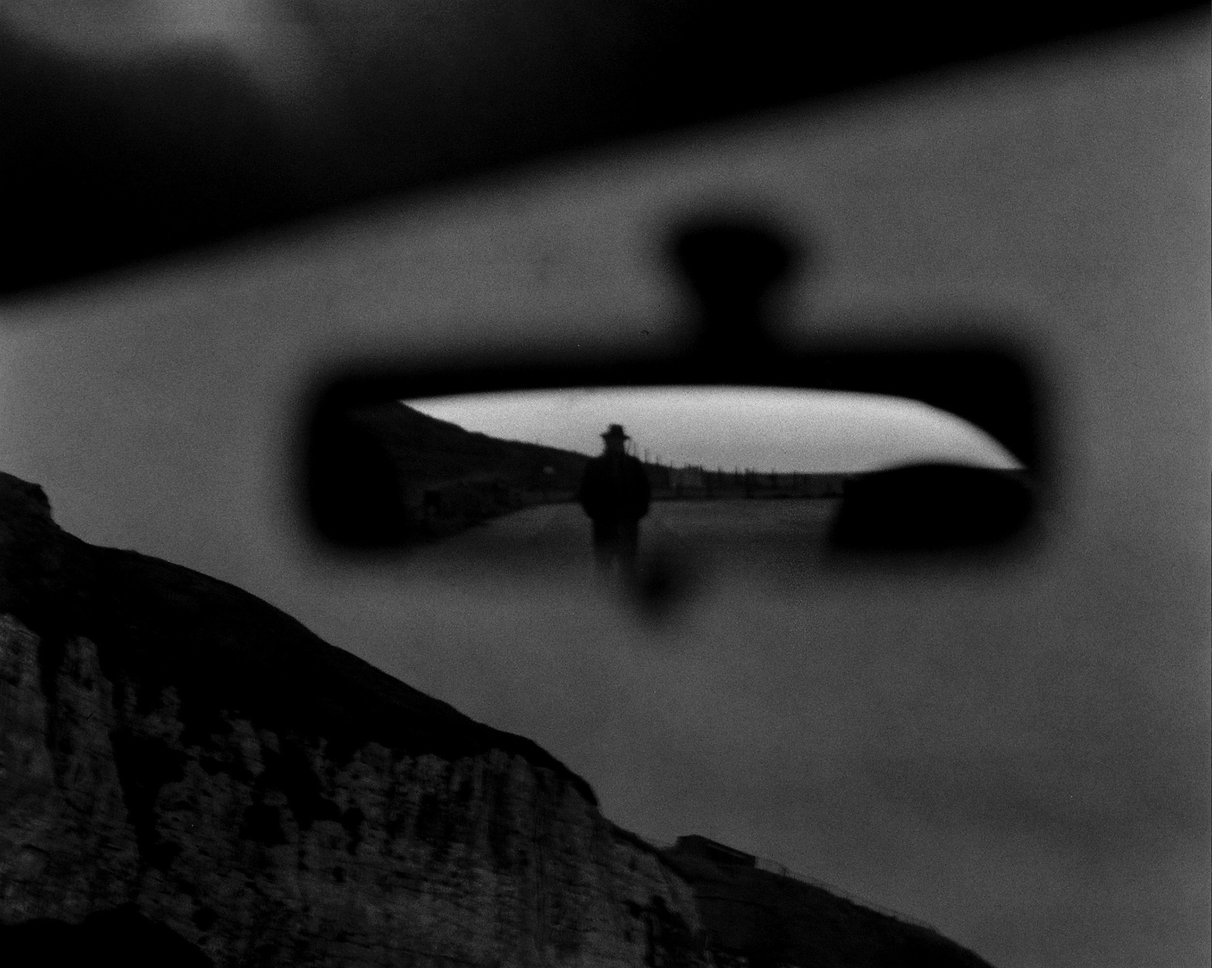 Silhouette of a man in a hat in a rearview mirror. The image is blurry except for the silhouette in the rearview.