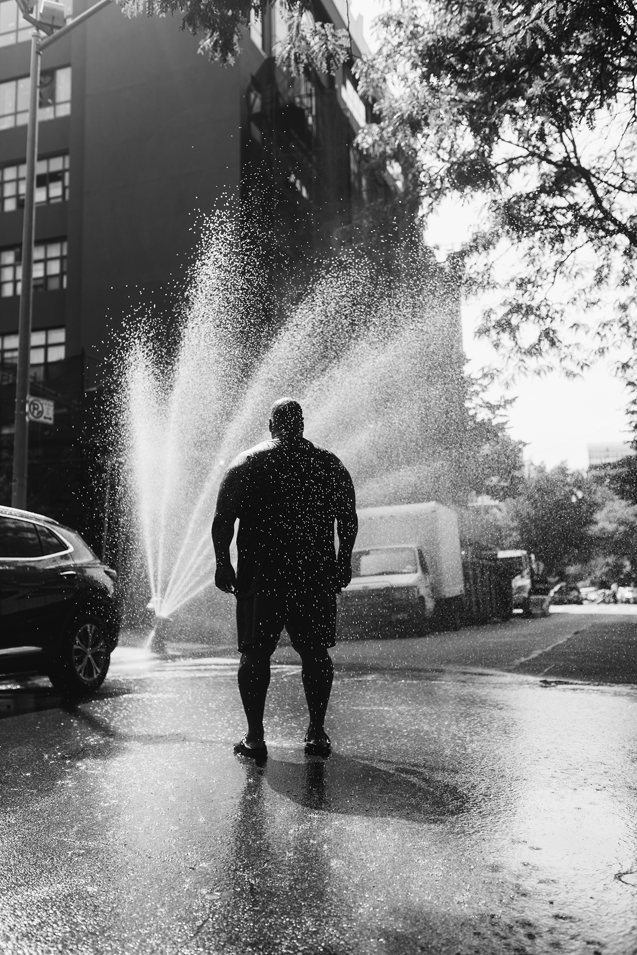 A large, muscular man seen from behind stands in the spray of water from an uncapped fire hydrant in Brooklyn, New York, on a hot summer day.