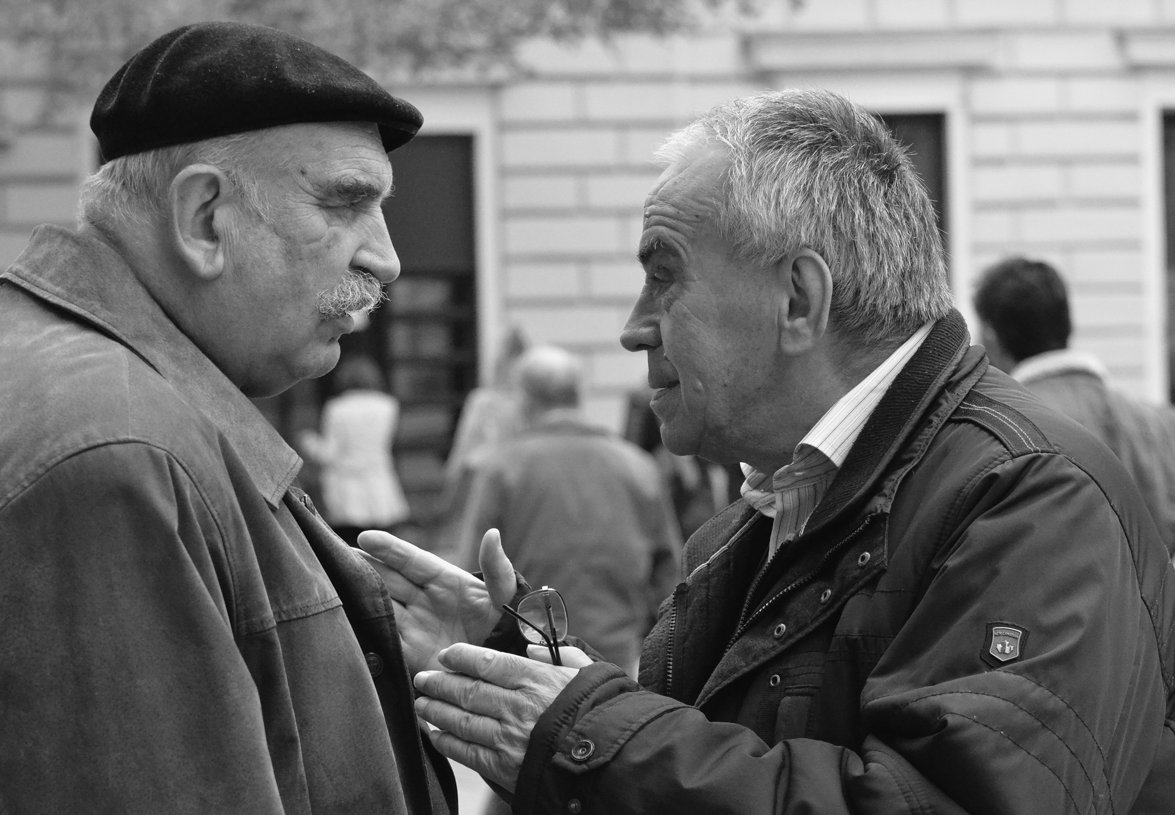 Two older men in jackets stand outside facing each other having an intense conversation in front of a building others are walking into.