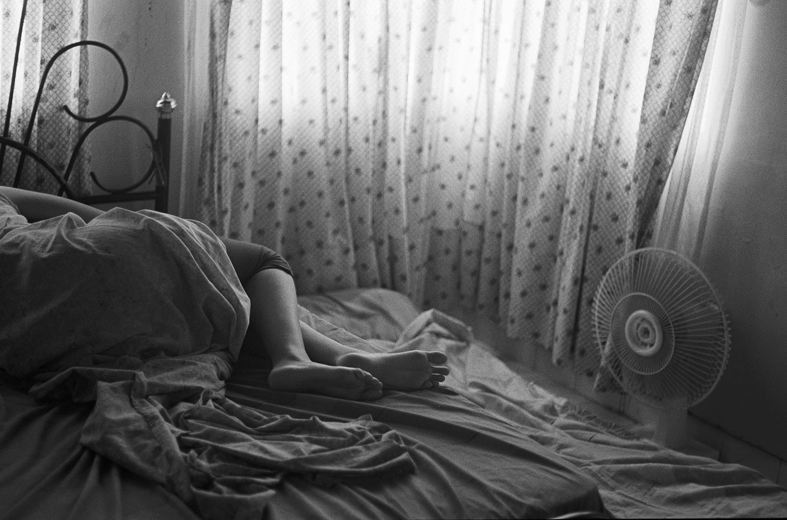 A person lies on a bed with their legs out from under the covers in a bedroom with sheer polka-dot curtains and a small fan on the floor.