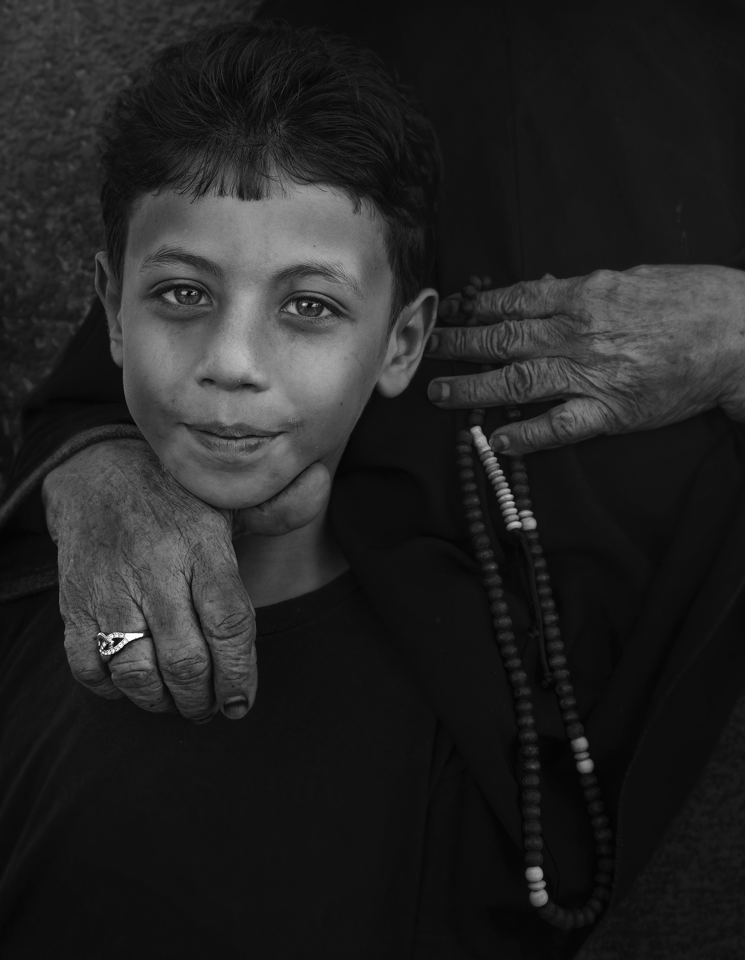 A boy looks at the camera as he stands in front of someone who has their right hand on the boy’s shoulder and holds a string of beads in their left hand.