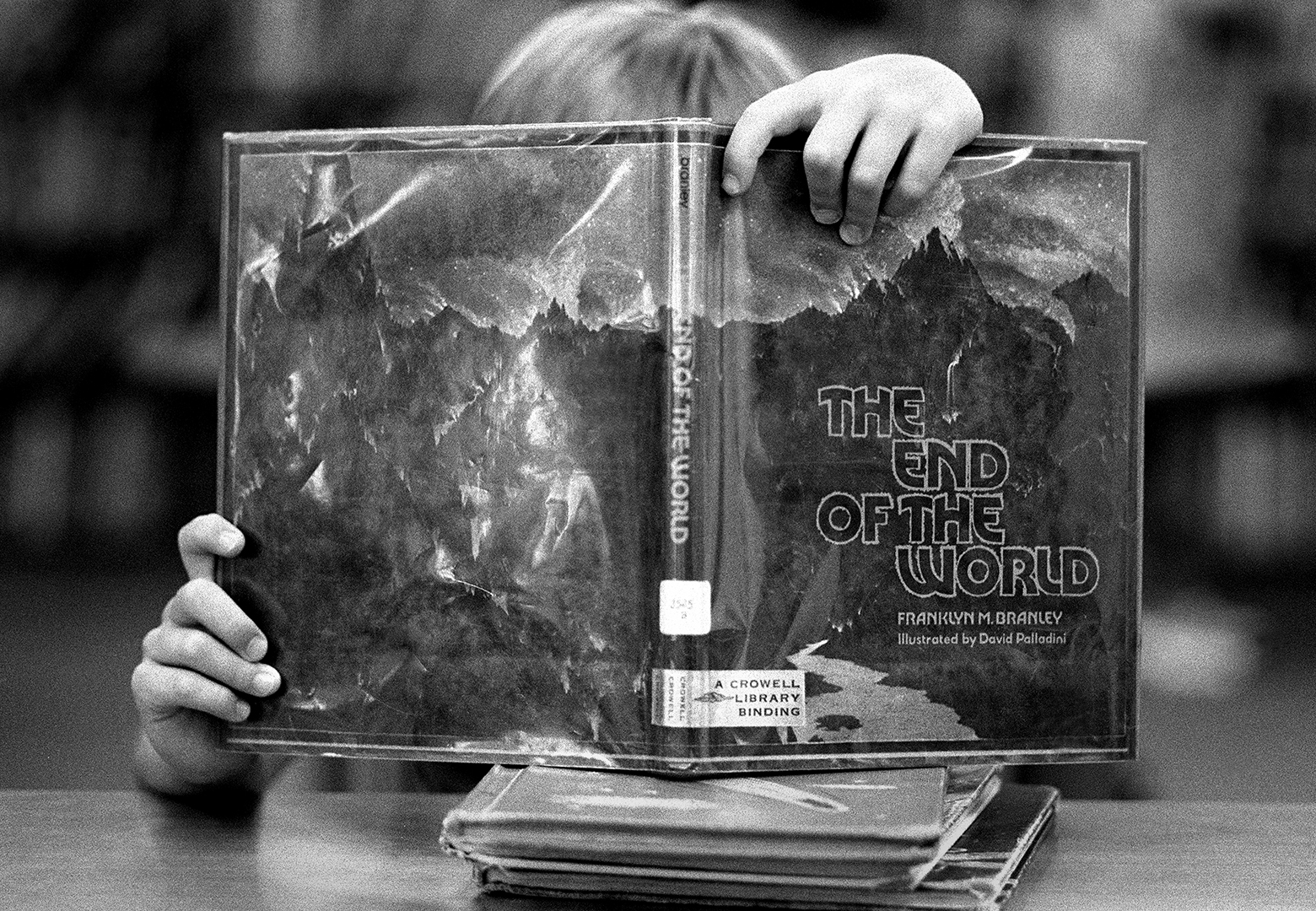 An elementary-school student sits at a table reading The End of the World in a library during a summer reading program. The student’s face is hidden behind the open book that is at eye level because they have perched it atop a few other books.