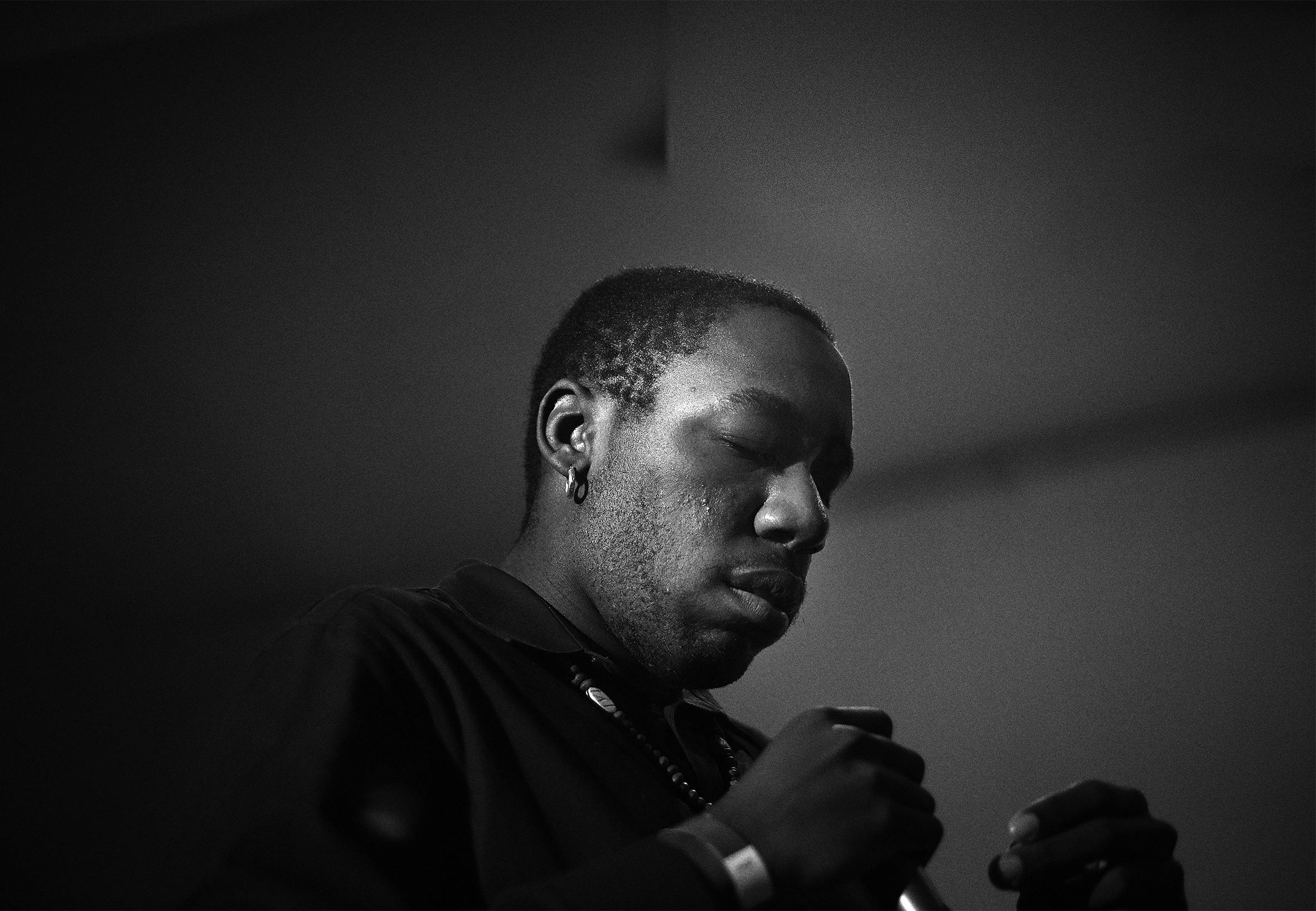 Close-up of a man from the chest up who has his eyes closed with his closed hands visible in front of his chest. He is wearing a collared shirt, necklace, earring, and bracelet.