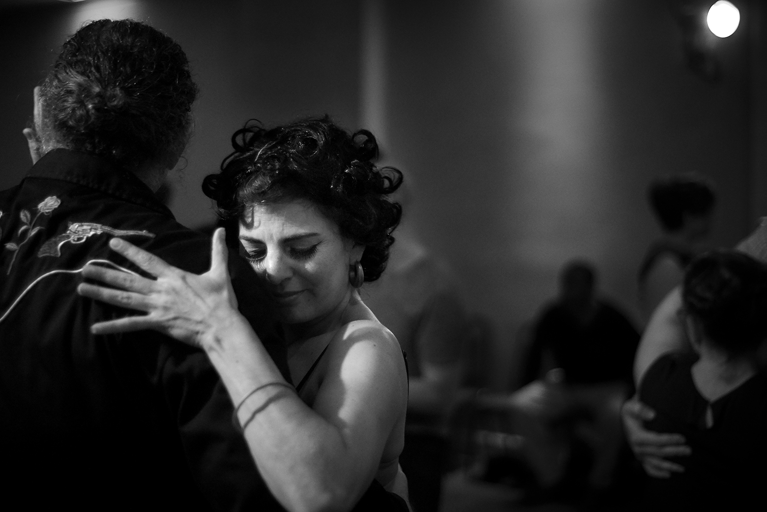 A man and a woman partner dance. He is taller than she is and seen from behind. She has her left hand flat on his back near his right shoulder. Her eyes are closed and she has an intense look on her face.