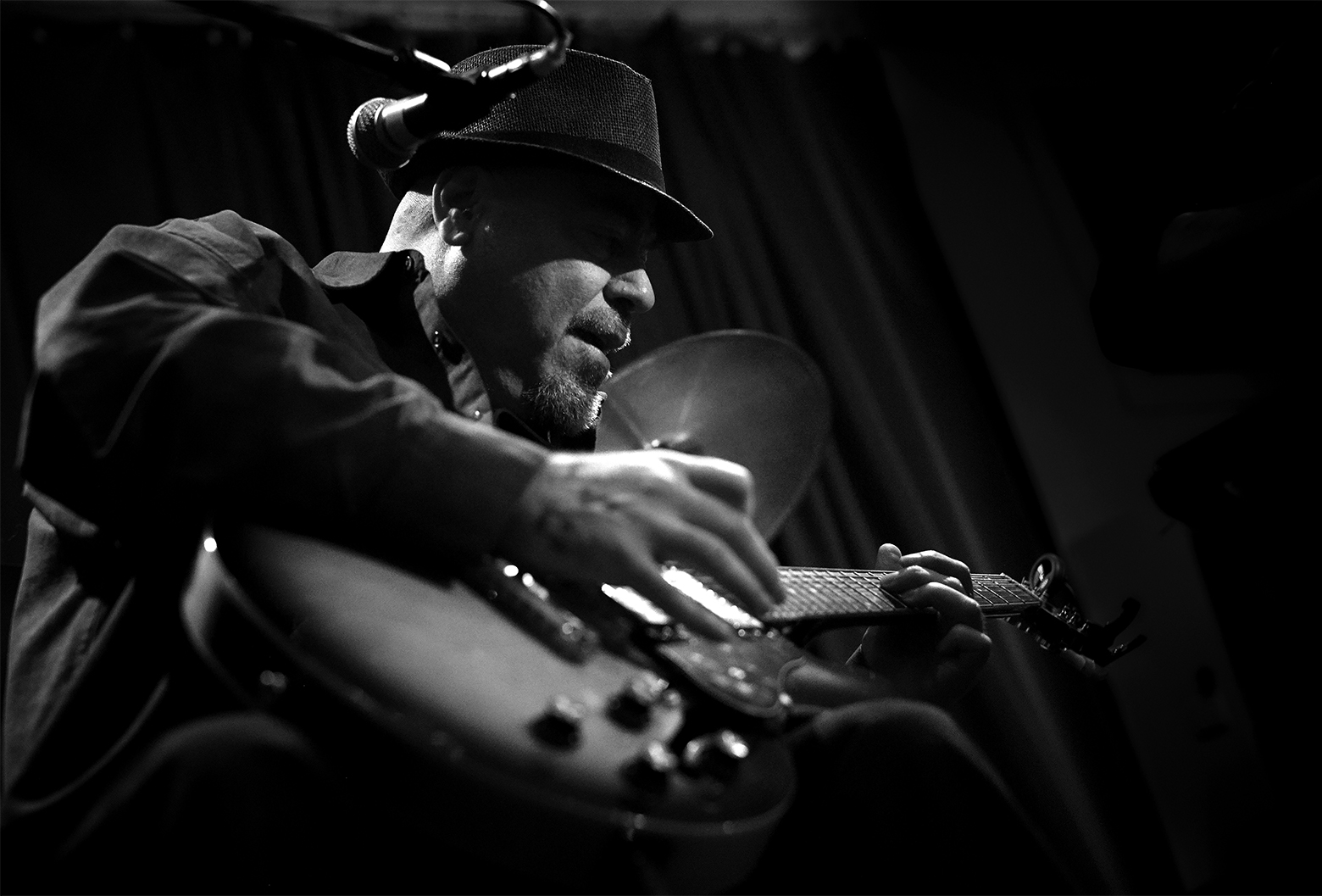 A man in a hat and collared shirt sits and plays the guitar.