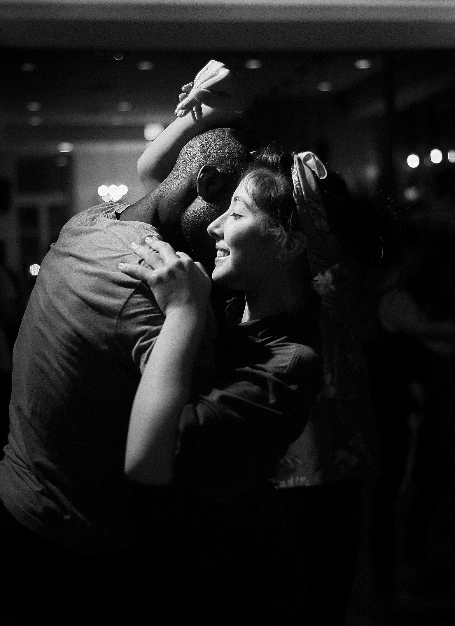 Side view of a man and woman partner dancing. His face is not visible. She smiles with her eyes closed with her left hand on his right shoulder as her right forearm rests against the side of his head while her fingers create a lovely shape above it.