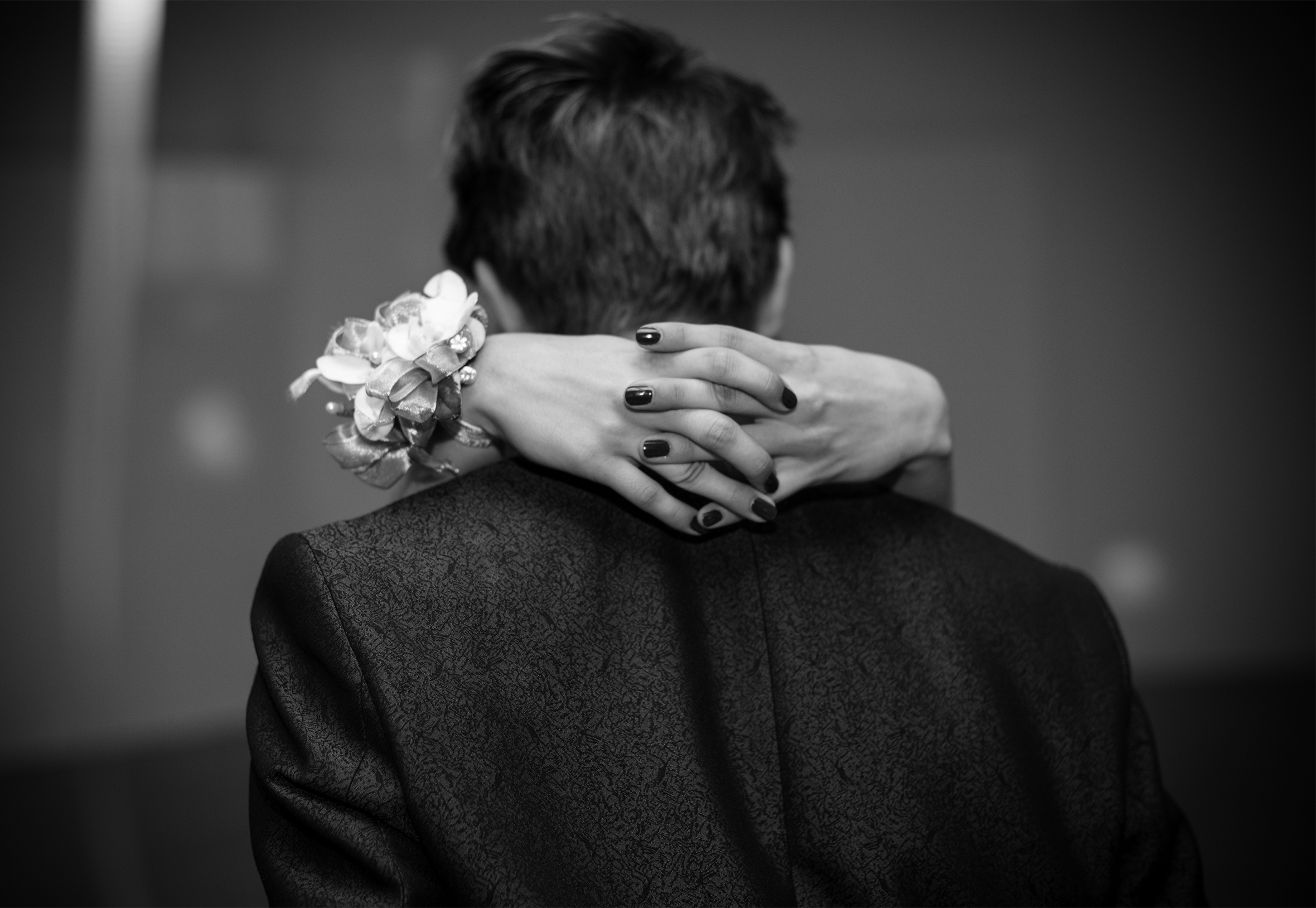 A couple slow dance at their senior prom. He is wearing a sports jacket and seen from behind. Of her, only her clasped hands around his neck resting on his shoulders are visible. She has dark fingernail polish and a corsage on her right wrist.