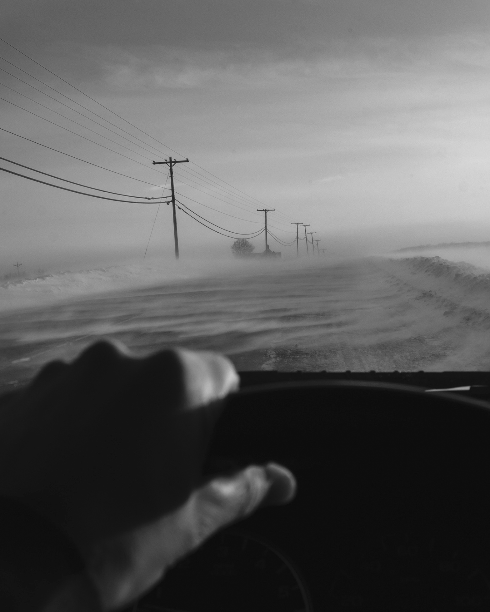 Close-up of a sunlit left hand on a steering wheel. Through the car's windshield utility poles are visible on the left side of the snowy road to the blurred horizon.