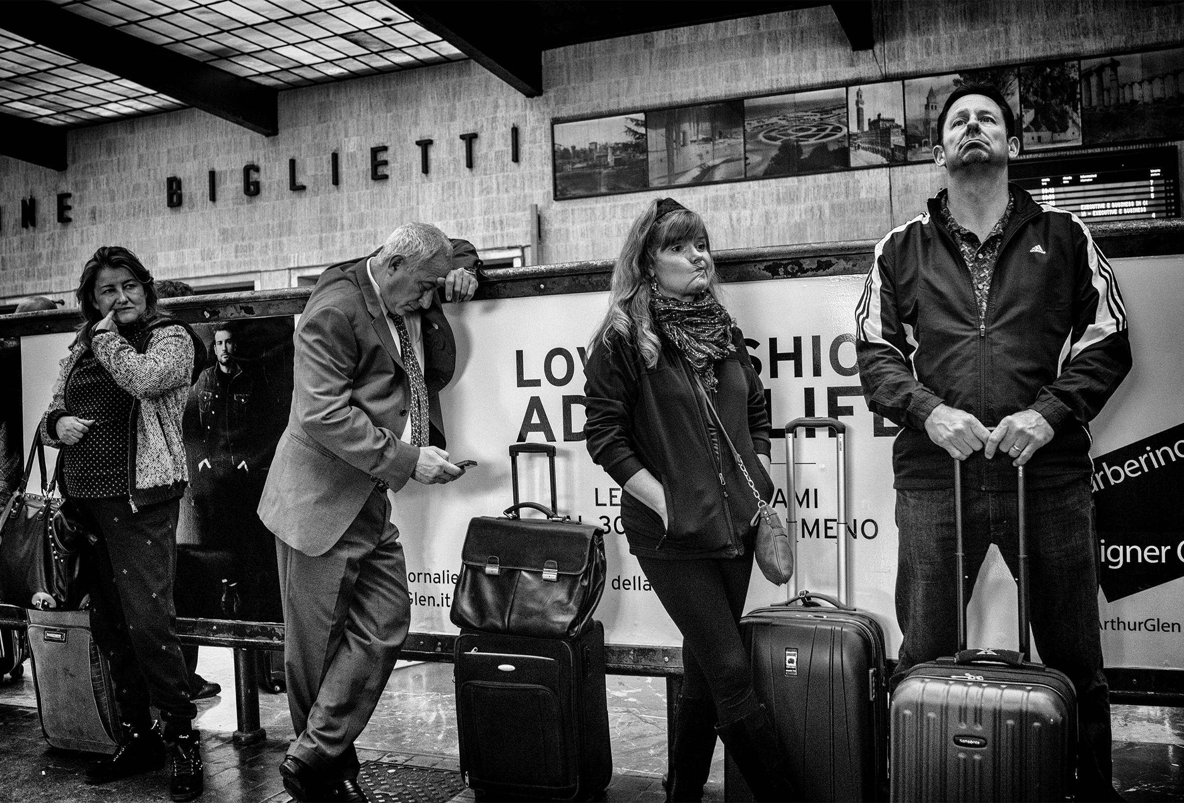 Four travelers at an airport in Rome stand and wait next to their wheeled luggage. From the left: A woman picks her teeth with her left pinky finger; a man looks down at his phone, a woman scrunches up her mouth; a man looks up while looking bored.