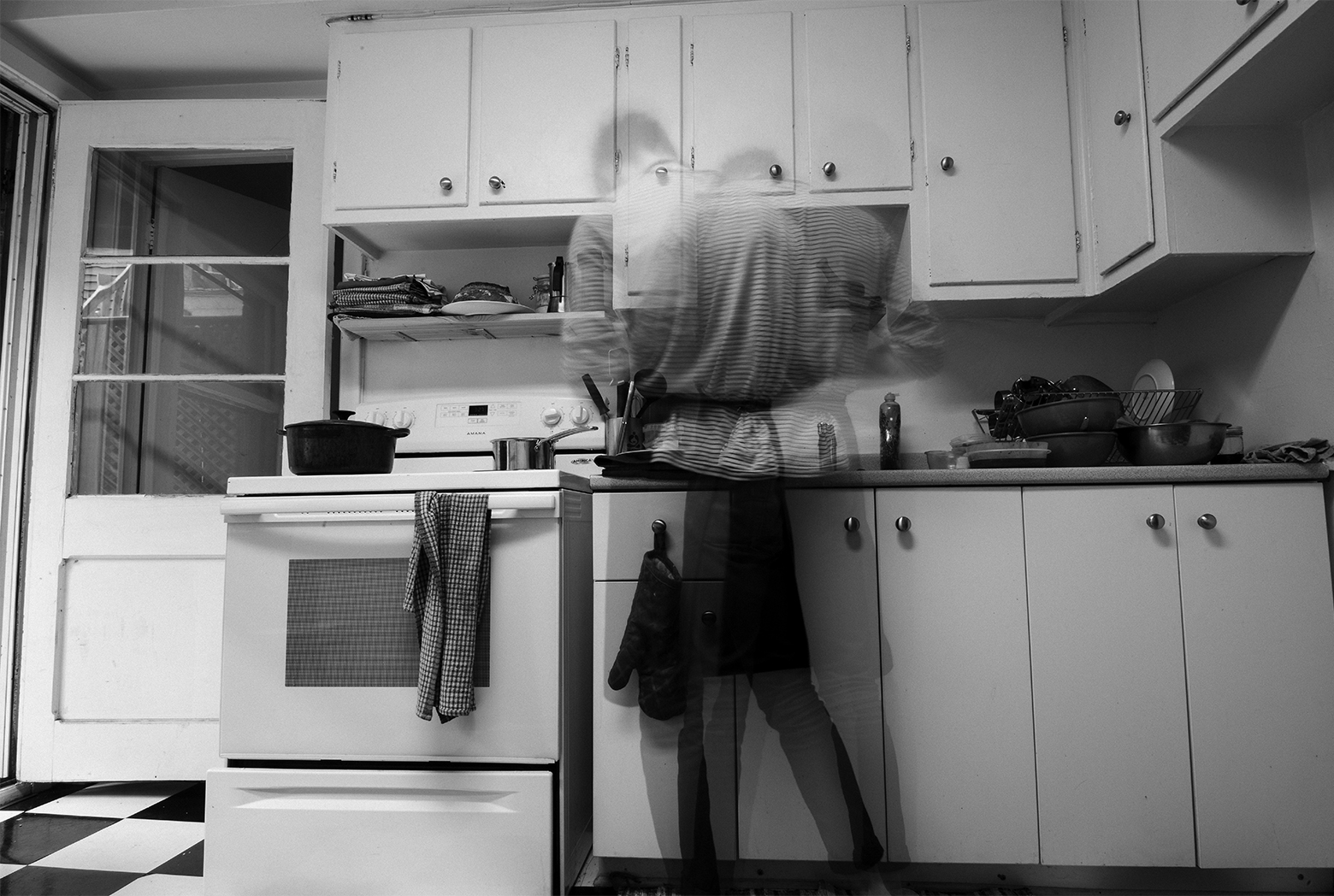 A man wearing a short-sleeved shirt and shorts cooks dinner in an apartment in Montreal with black-and-white checked flooring and white cabinetry. He is blurry and seen from the back with several images seemingly superimposed to depict his movement.