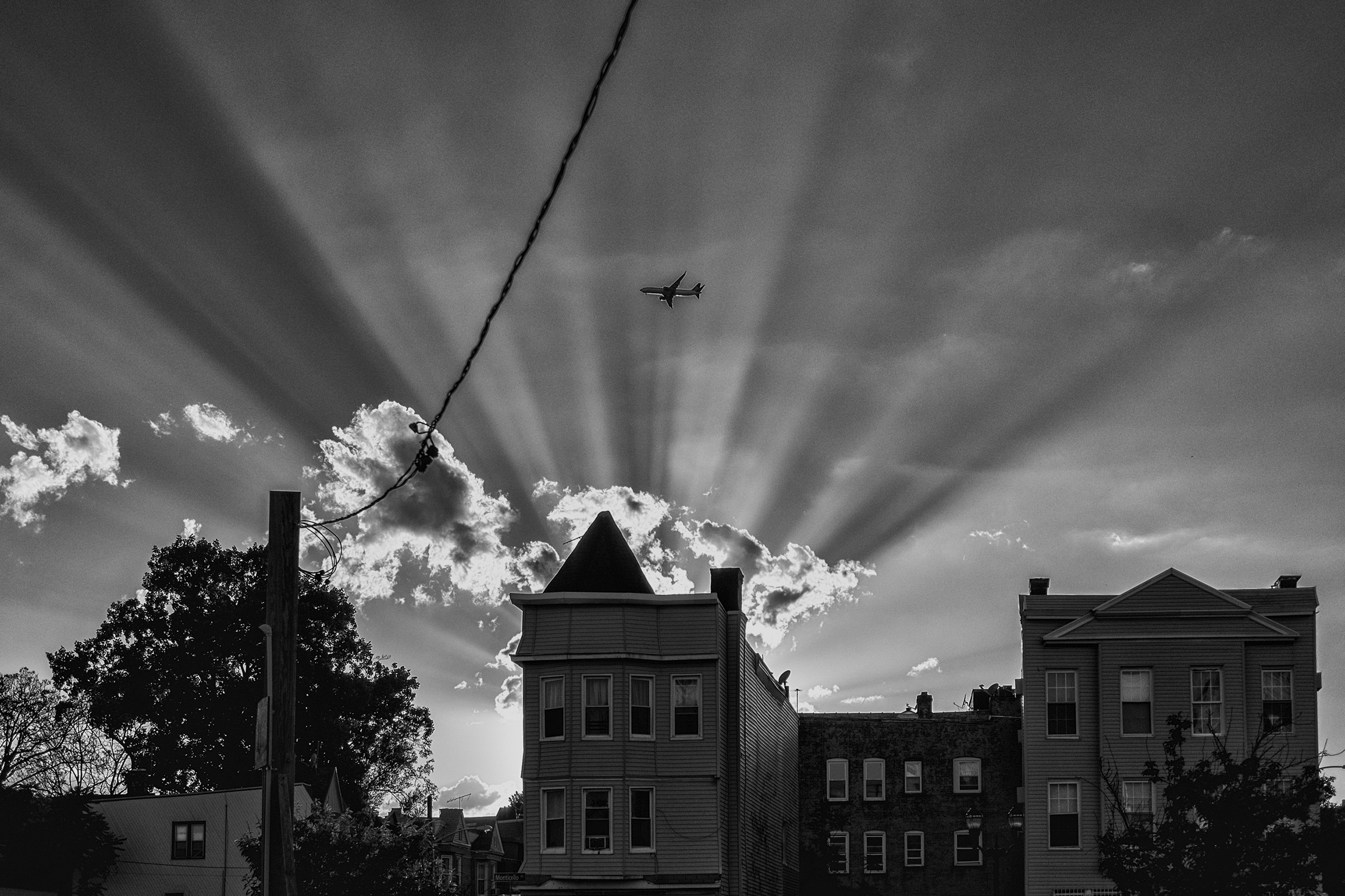 Older apartment buildings with a telephone pole on the left are in front of a grouping of clouds backlit by the sun. A plane traveling to the left is amid a burst of dark shadows going from the grouping of clouds to the top of the image.