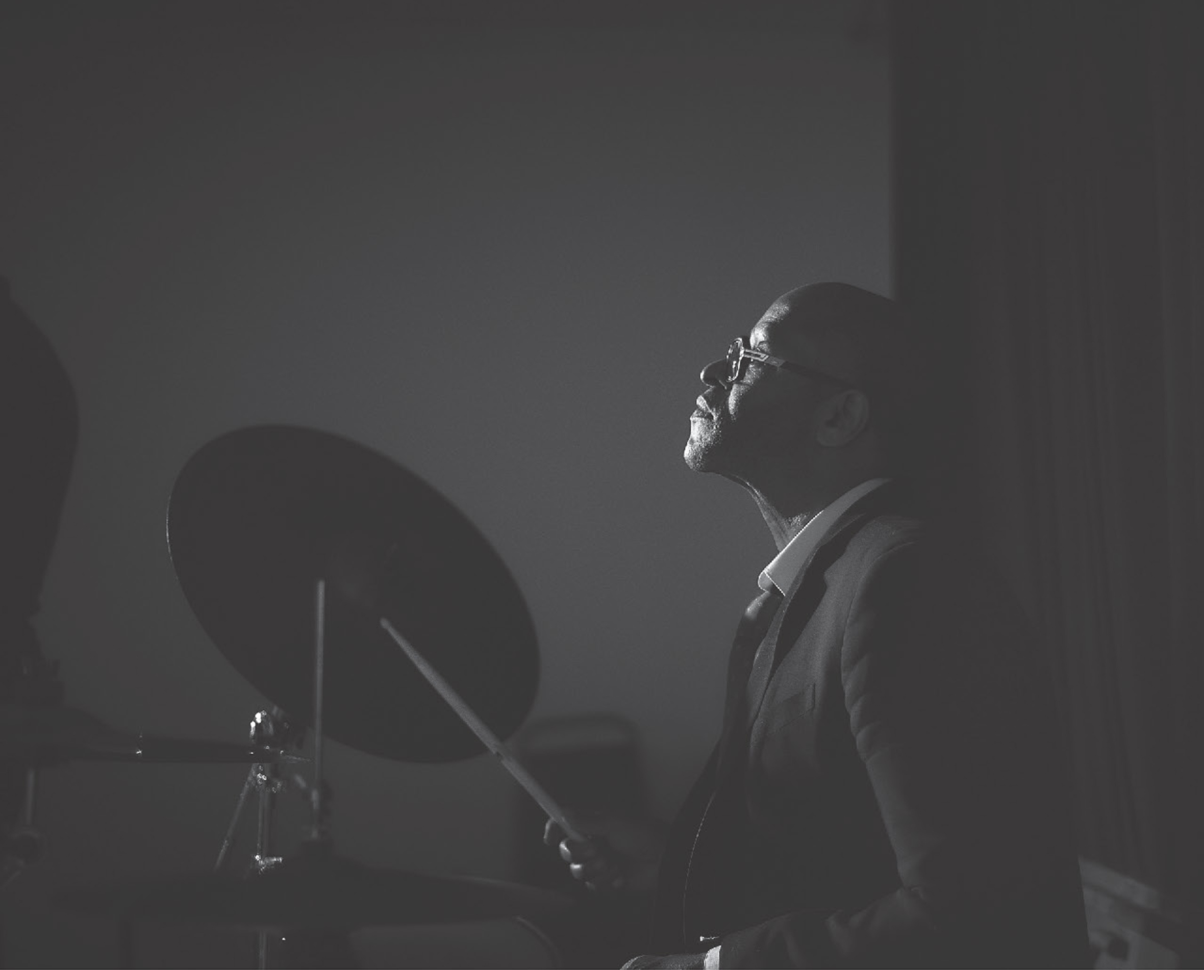 A man in a suit coat, shirt, and tie looks ahead as he sits and drums.