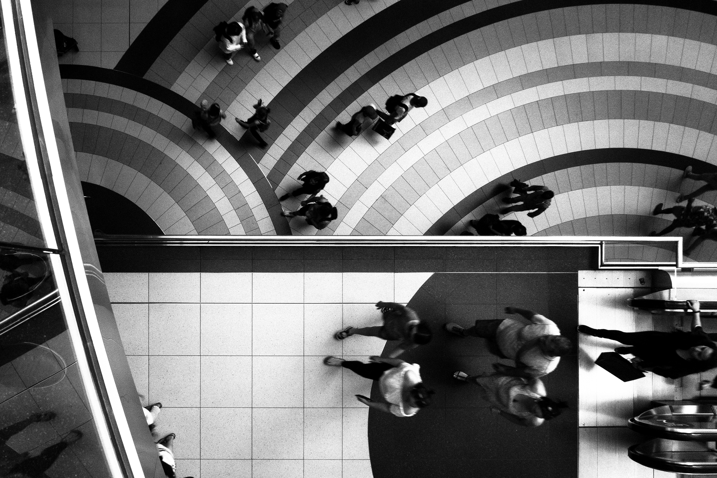 Many people walking in a Toronto mall. There are three levels of walkways visible: the top and bottom have square tiles; the middle has rectangular and square tiles set in circular patterns sporting multiple stripes of varying widths and color tones.