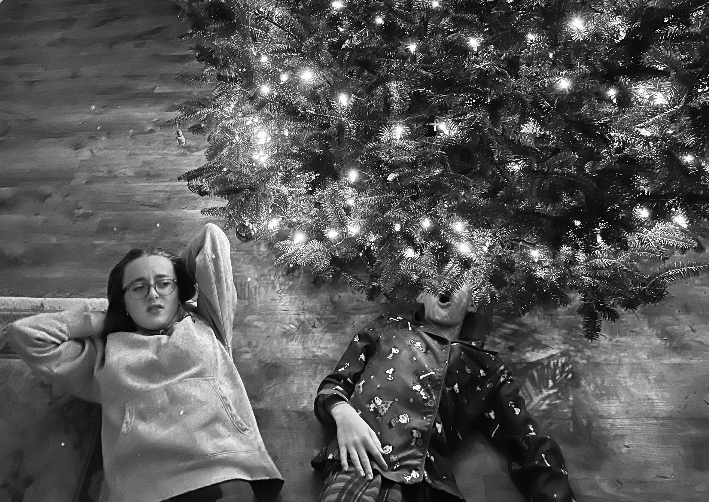 A sister and brother lie on their backs for a new perspective on the Christmas tree that was just strung with lights. She looks disinterested. He has positioned his face underneath the tree and it is not visible except for the O-shape of his mouth.