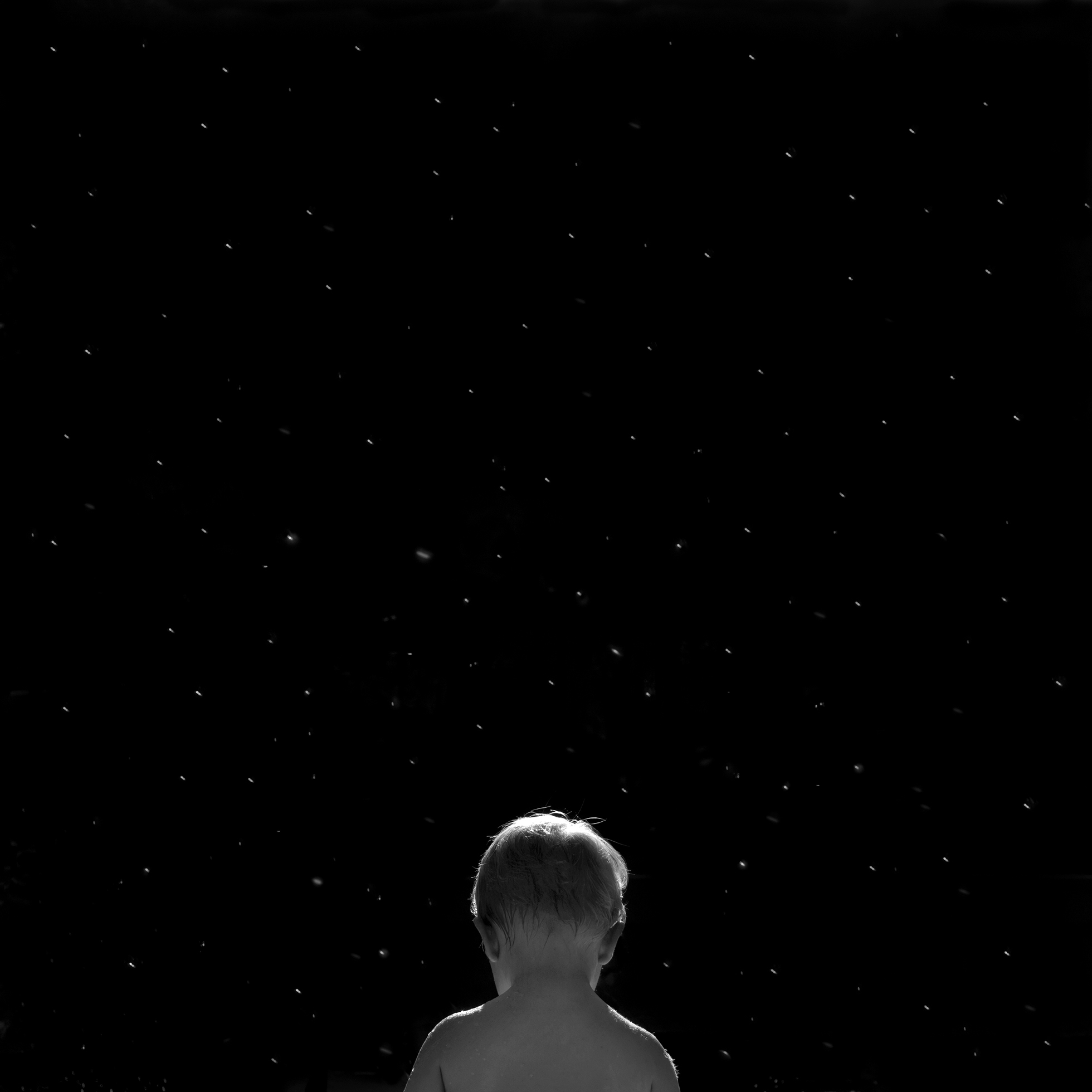 A blond boy seen from midback up and from behind looks down as he is surrounded in the black night by a sky full of stars.