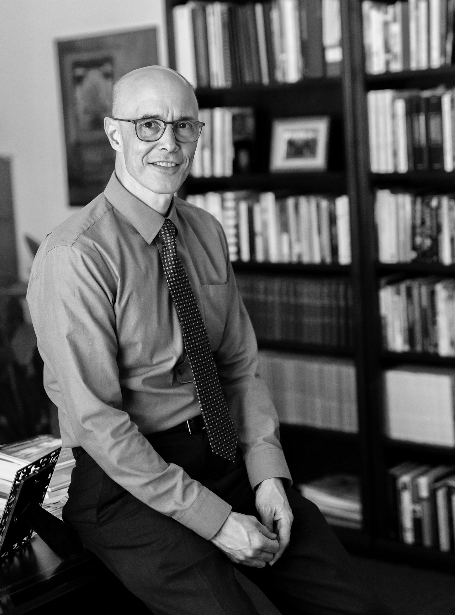 Jim Tucker smiles as he sits on the edge of a desk with his hands clasped in front of him on his thighs. A very large, very full bookcase is behind him and to his right. He is wearing a long-sleeve shirt with a tie and slacks.