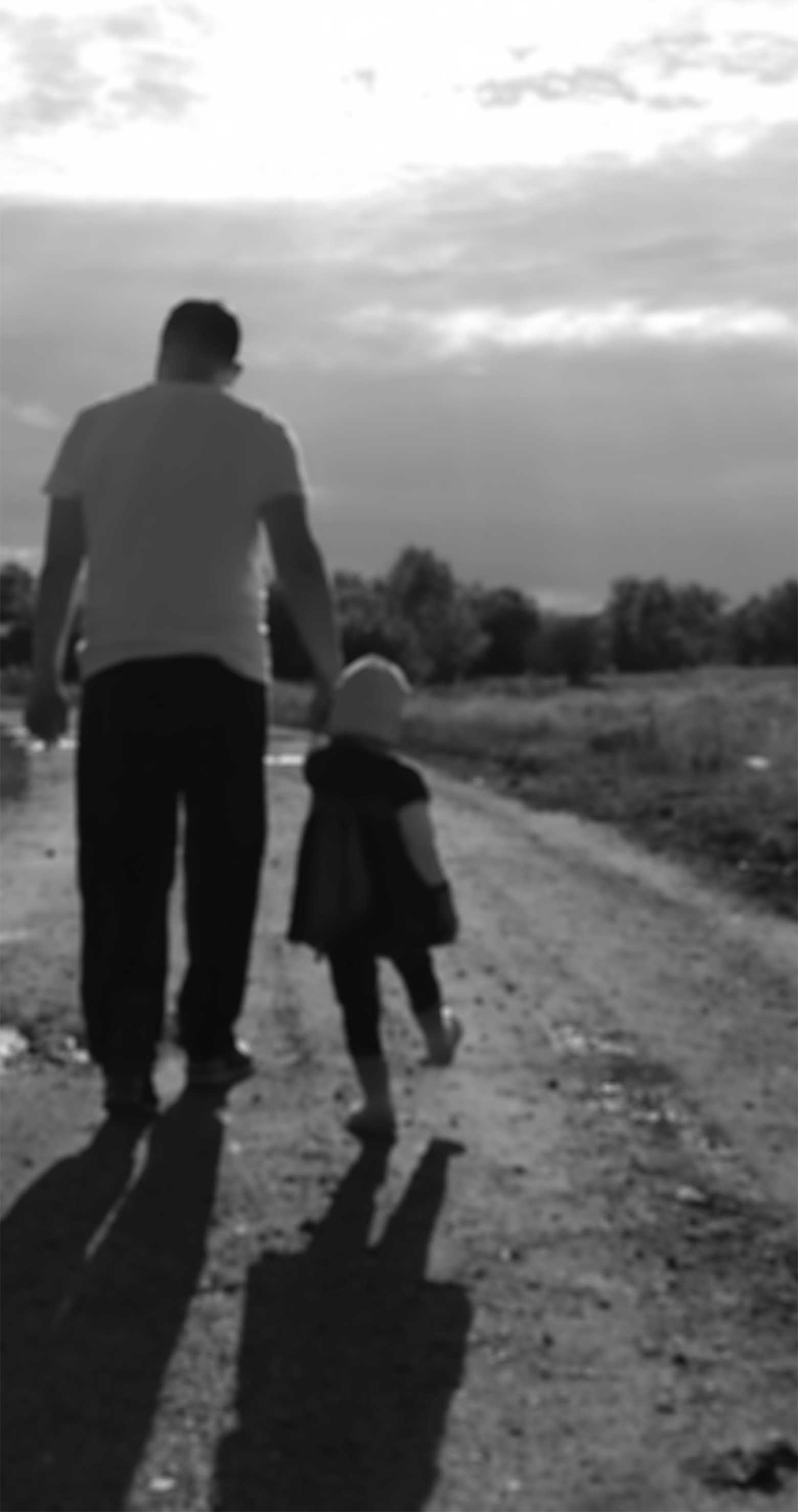 A father and young daughter take a walk on an unpaved road, with trees in the distance, and enjoy the sunset near their home. He is wearing a white T-shirt and dark pants; she is wearing a light-colored hat and boots with a long top and leggings.