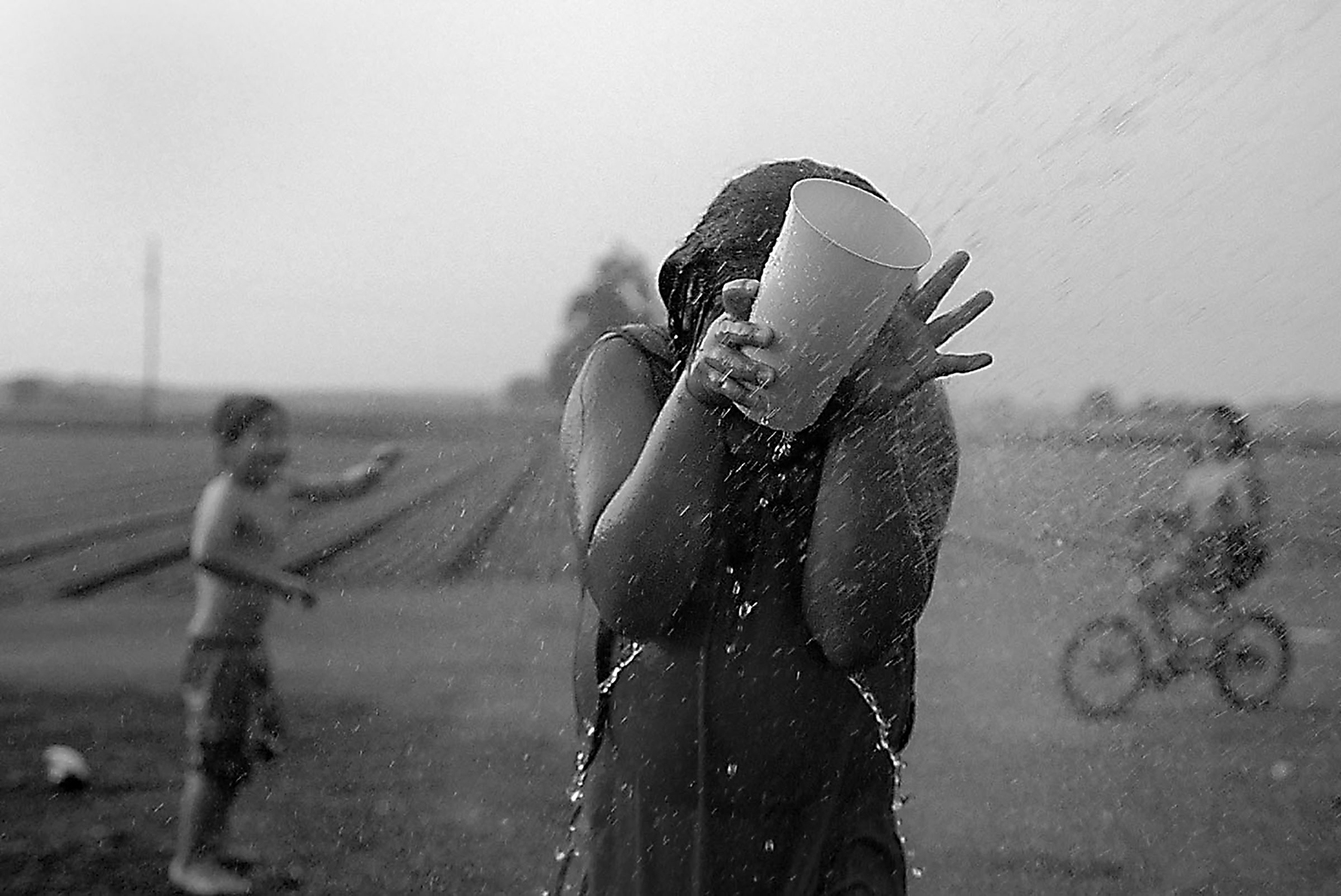 A young girl stands soaking wet holding a large cup in front of her face as she is being squirted with water on a warm summer day in Hartville, Ohio. Behind her to the left is a young boy and to the right is a child on a bicycle.