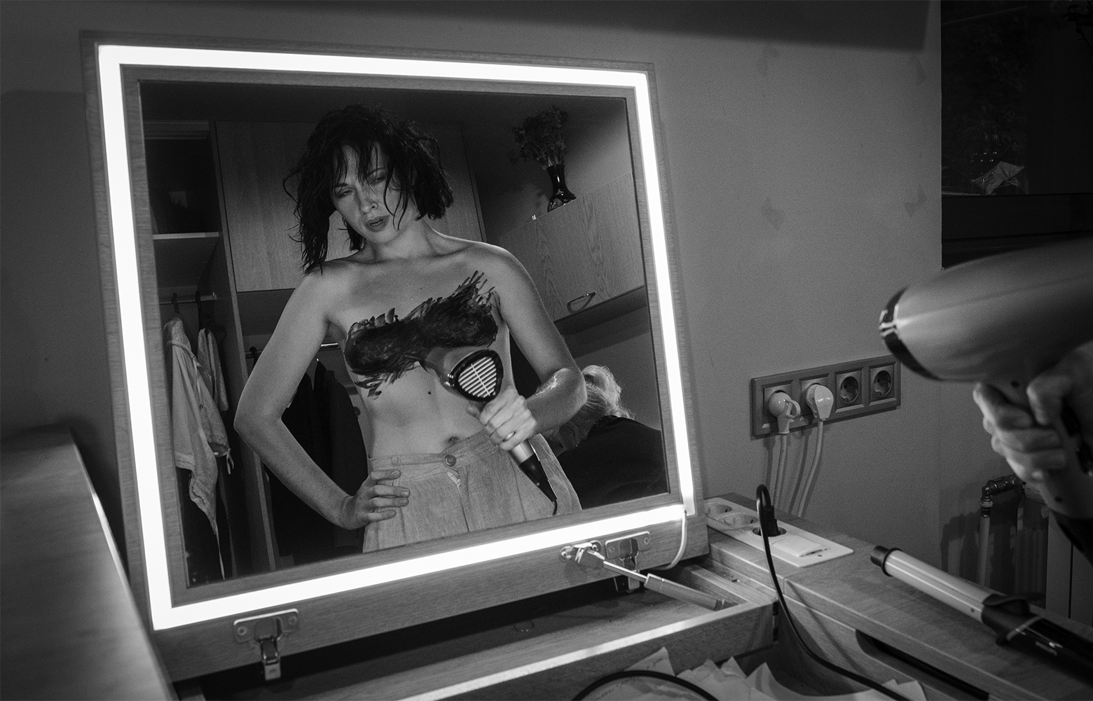 Woman viewed in a mirror in a dressing room backstage as she blow-dries makeup covering her breasts. She is nude except for a pair of pants.