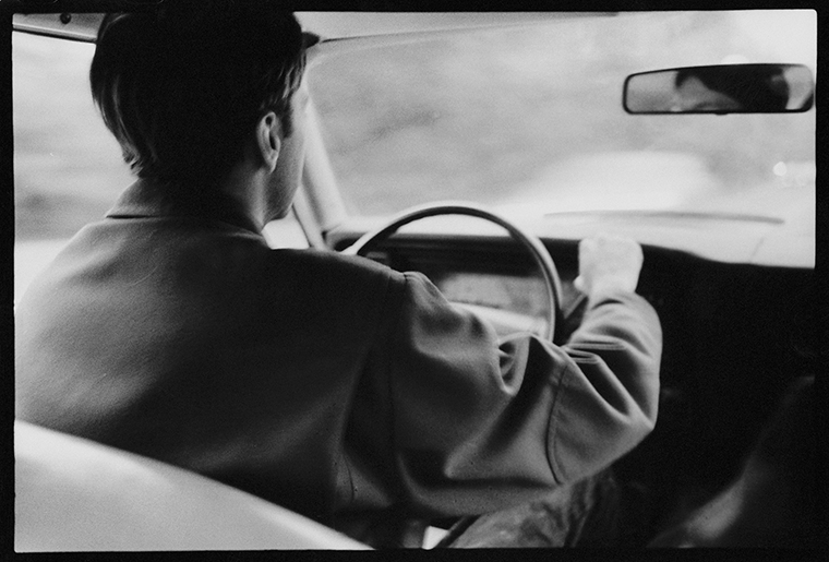 A man wearing a jacket and seen from the back seat is driving an older model car.