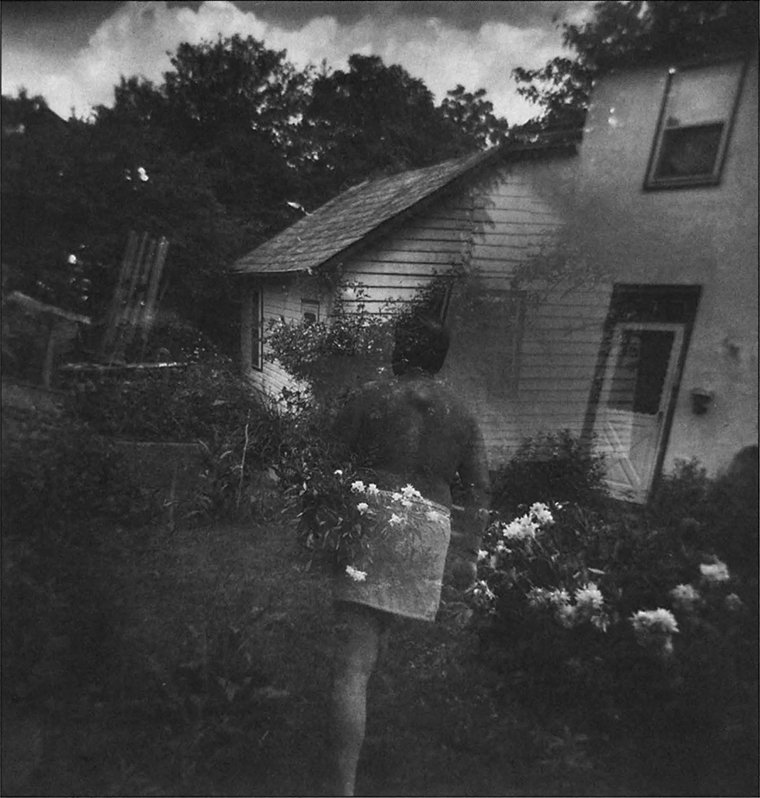 Blurry image of a nude man from behind with a towel wrapped at his waist walking through a yard full of foliage next to a house that is off-kilter.