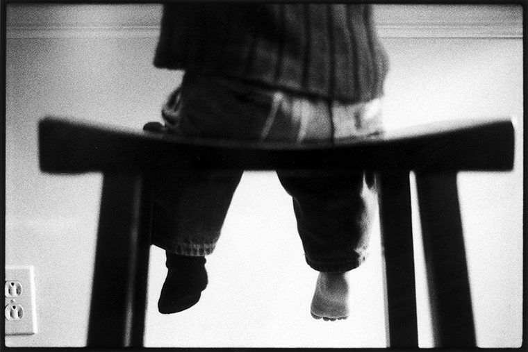 A barefoot toddler wearing a shirt and jeans is seen from the waist down and from behind sitting on a stool.