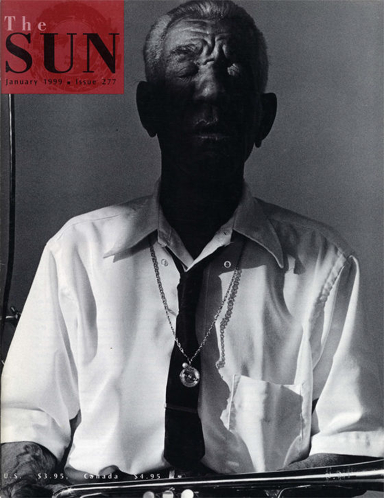 An old man with his eyes closed in a white button-down short-sleeve shirt and tie with a necklace on top sits with a musical instrument on his lap.