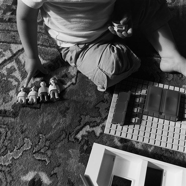 A young child sits on a carpeted floor with plastic figures of a man, woman, and two girls as well as household furniture ready to snap on to the gridded floor of a toy model.