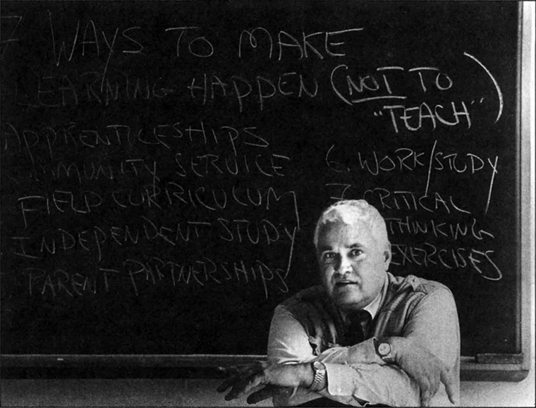 John Taylor Gatto sits facing the camera with his arms casually crossed. The blackboard behind him says 7 Ways to Make Learning Happen (Not to Teach).