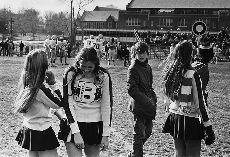 Two high-school cheerleaders on the sideline sharing a laugh as another teen looks over at them.