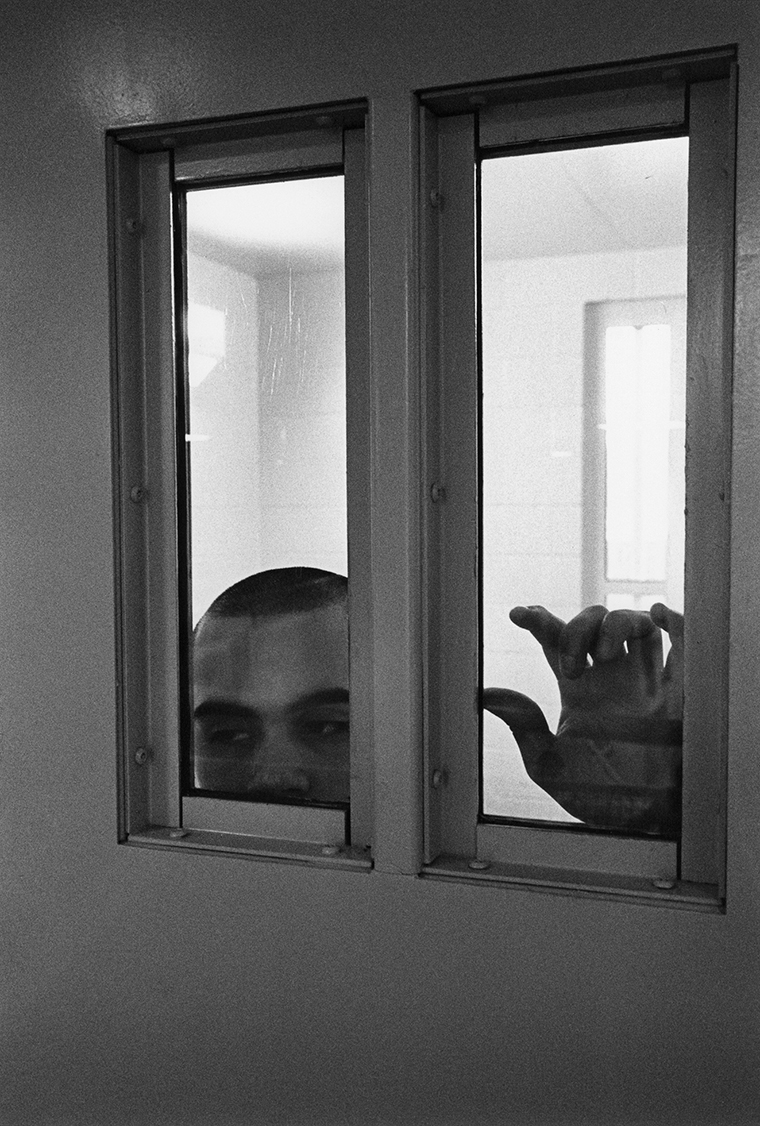 Edward, age seventeen, stands behind a metal door with two glass pane insets in San Jose Juvenile Hall’s maximum-security B-8 Unit for violent and high-risk youths. The top of his head and his palm on the glass are visible.