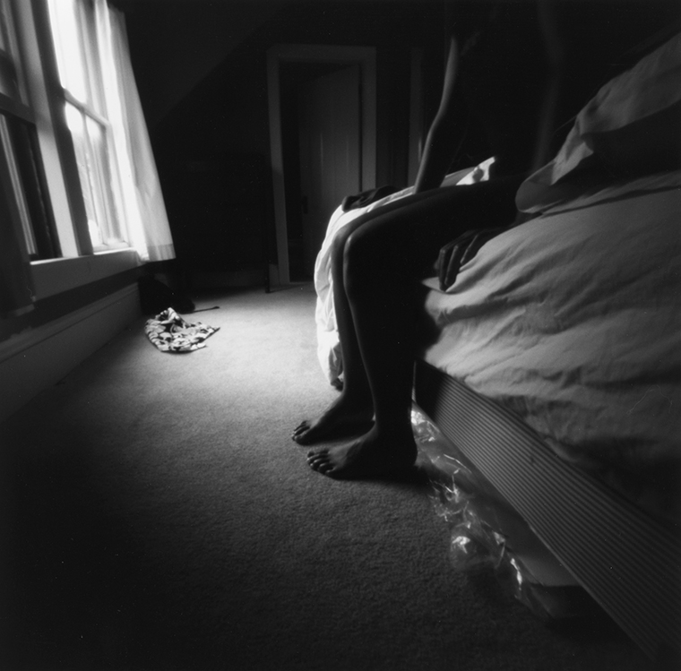 A woman in shadow sits on the edge of a rumpled bed.