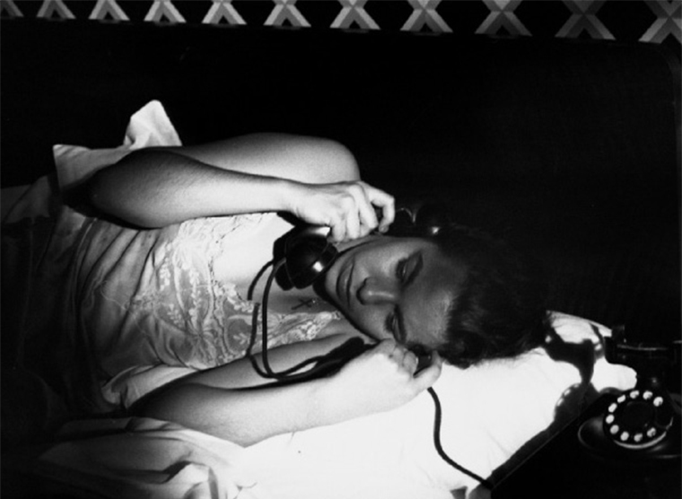 A woman lies on her side in bed with the handset of a rotary phone pressed to her ear.