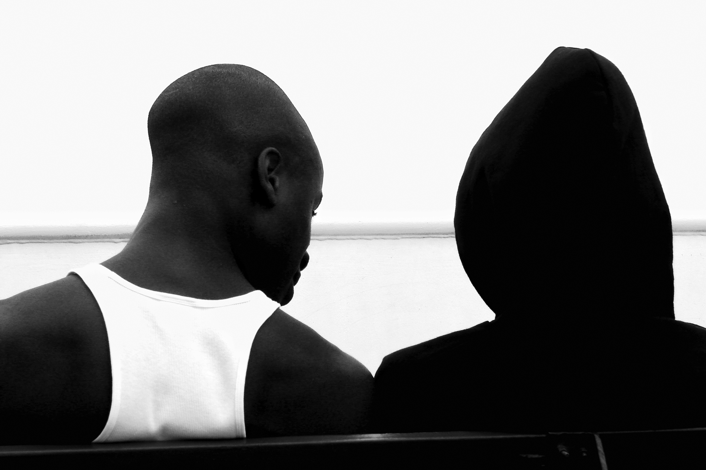 A young, fit man with a shaved head in a stringer tank top and a person in a hoodie with the hood up are seen from the back sitting on a bench with their shoulders touching on a ferry off Long Island, New York.