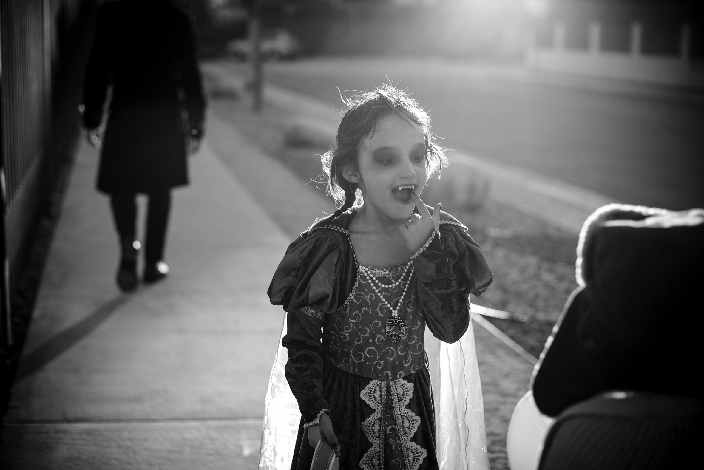 A little girl on a sidewalk dressed in a princess costume on Halloween is showing off her vampire fangs to a child with their back to the camera in the foreground.