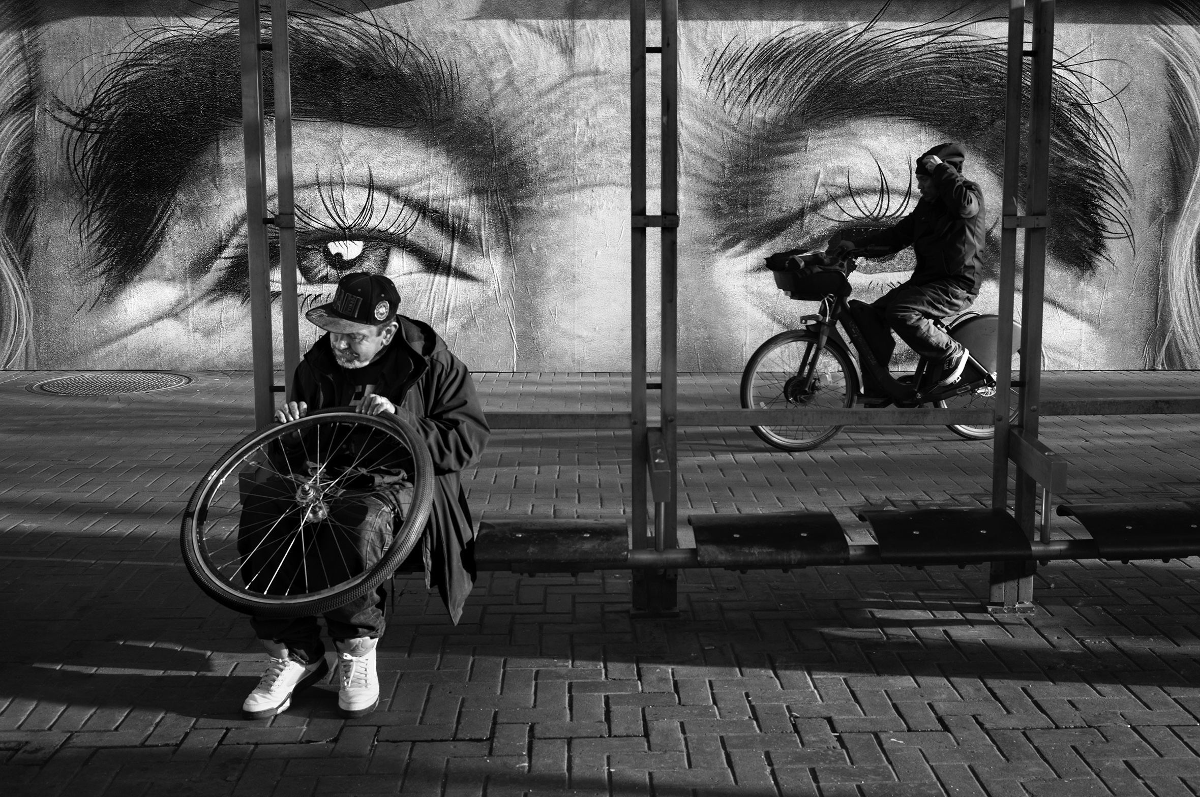 A man sits at a bus stop holding a bicycle wheel on his lap as another man rides a bicycle down the street behind him. The building behind the bus stop has a large, close-up image of a person’s eyes and bushy eyebrows.