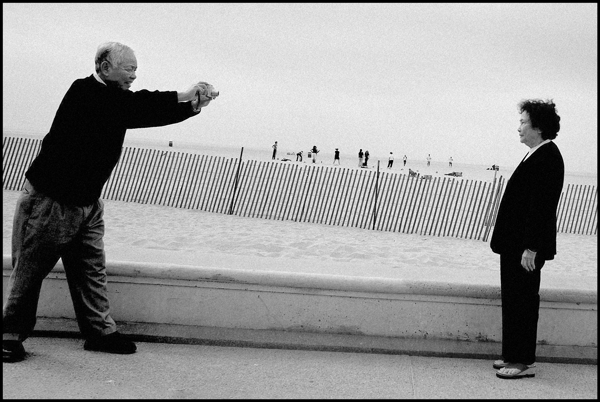 A man fully outstretches his arms and uses a small camera with lens to photograph his wife as she stands with her arms to her sides on a public boardwalk in Santa Monica, California.