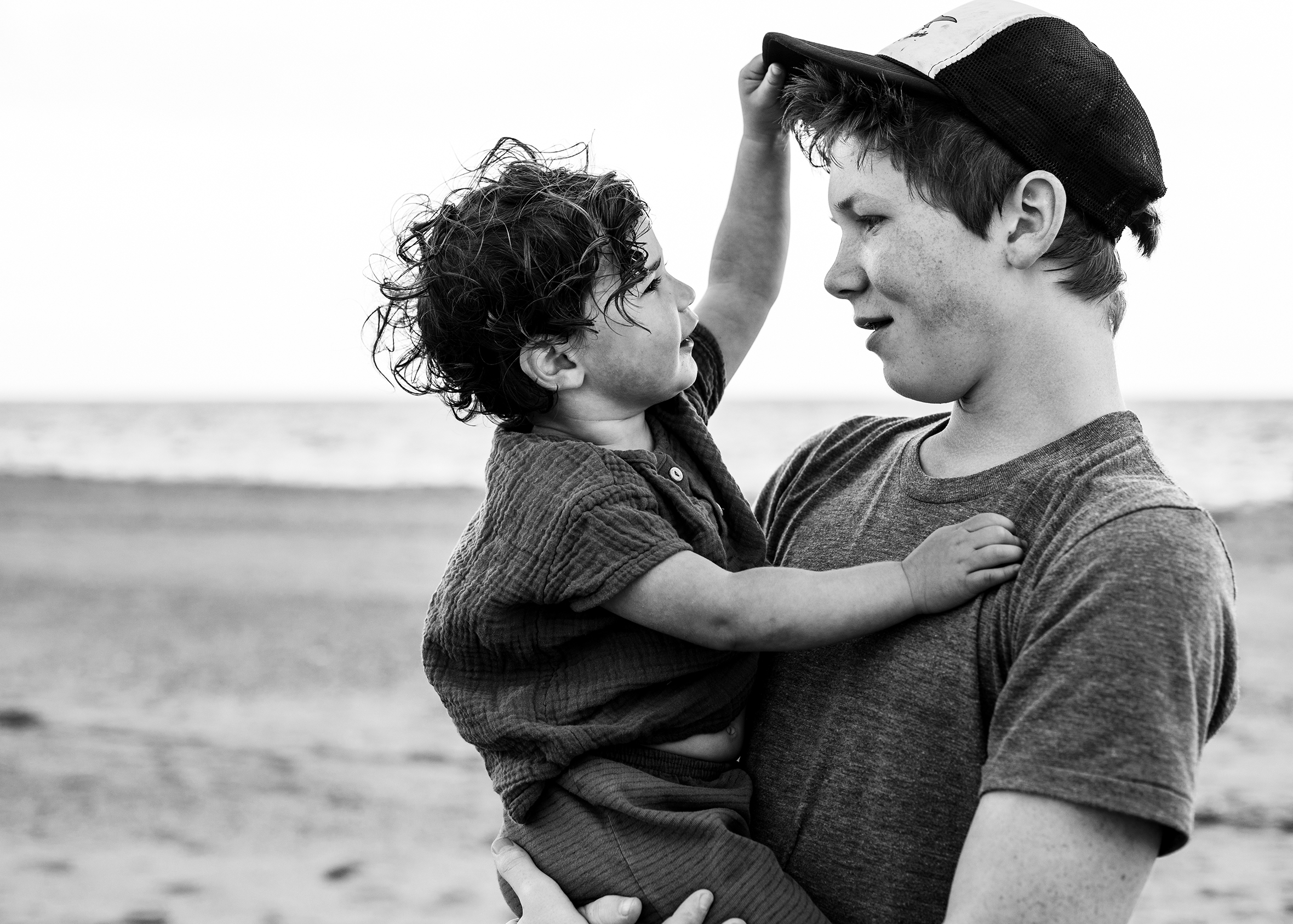 A teenager holds a toddler on the beach as they look at each other and smile. The toddler has curly hair and is touching her brother’s ball cap.