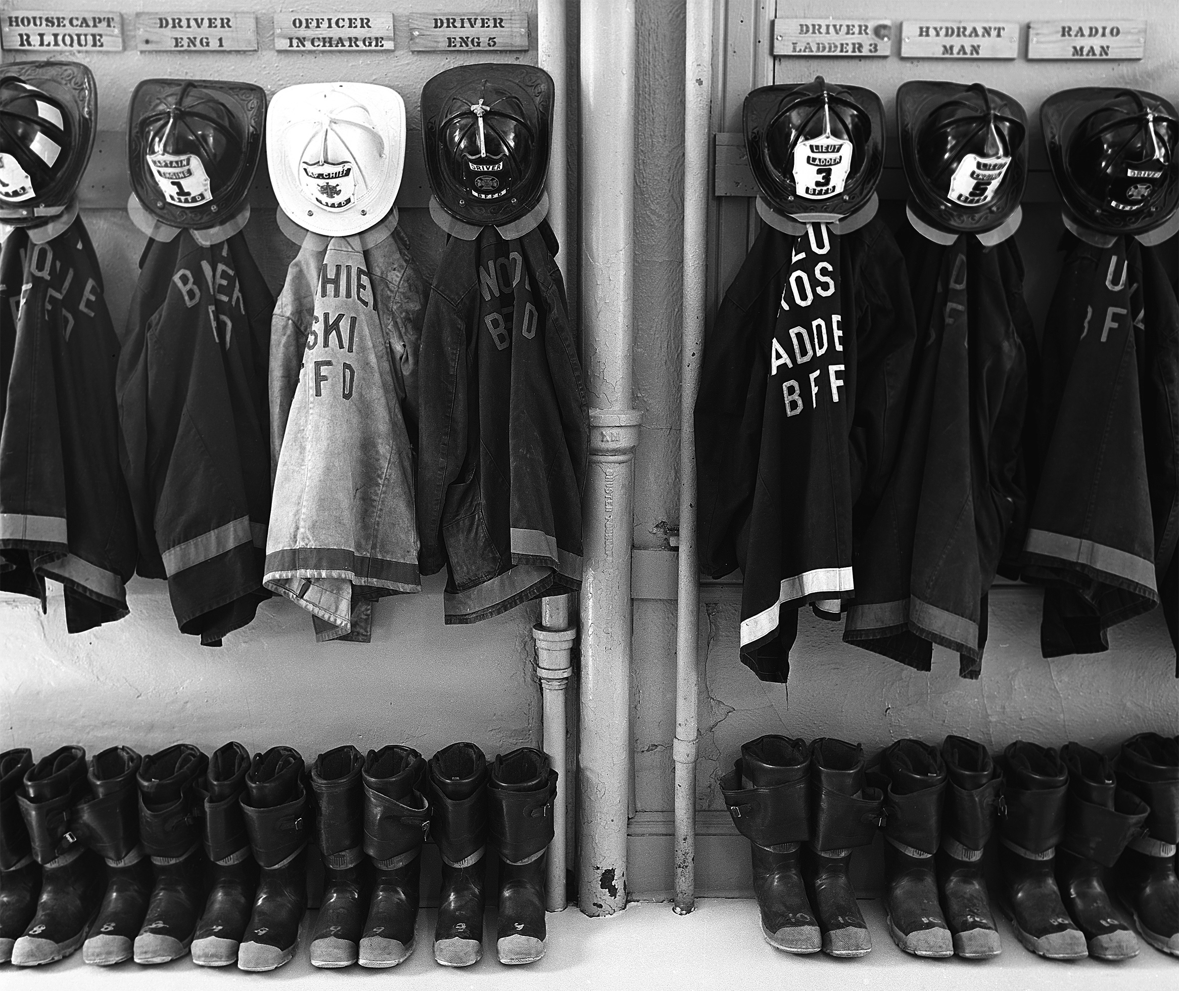 Firefighter turnout gear—helmet and coat—hang from a wall with boots aligned on the floor below in a fire station in Bellows Falls, New Hampshire, in 1978. Above each set of gear is a wooden plaque with an assigned task stenciled on it.