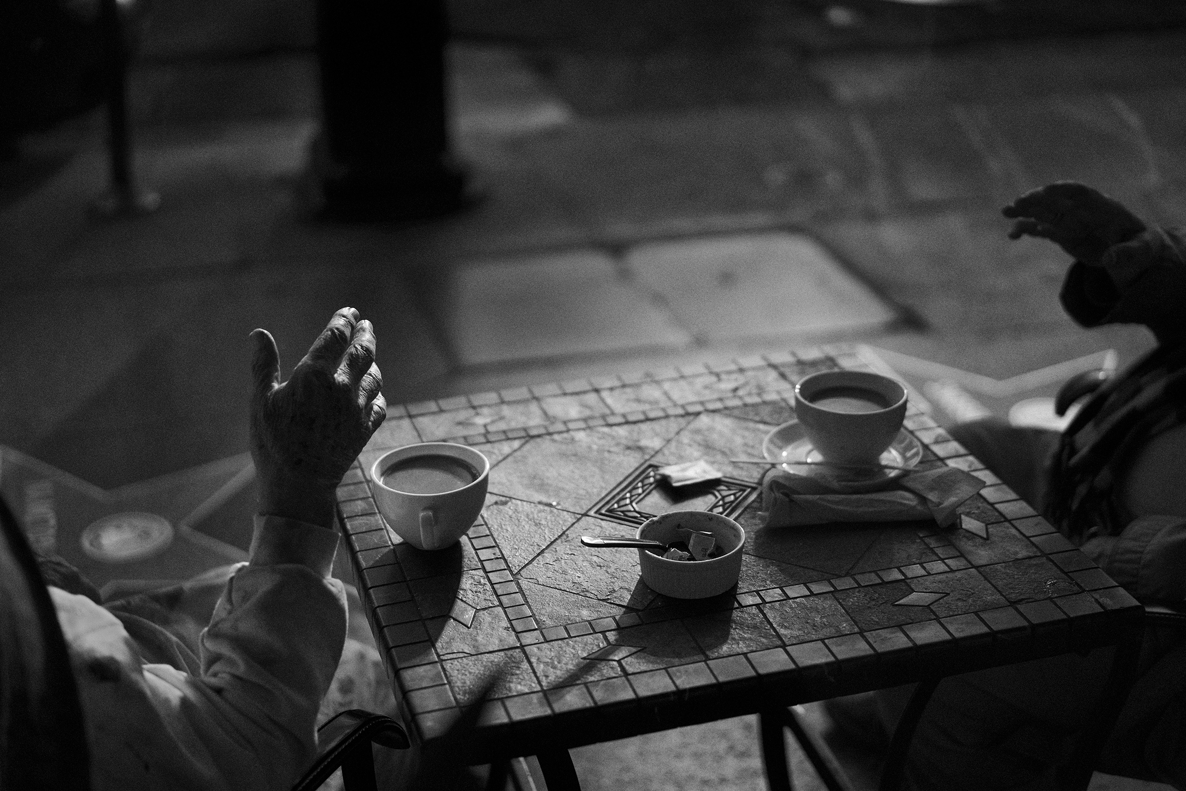 Two people turned toward each other sit at a small, square patio table with two full coffee cups upon it. They are in easy conversation evidenced by their relaxed hand gestures which is mostly all that is visible of them.