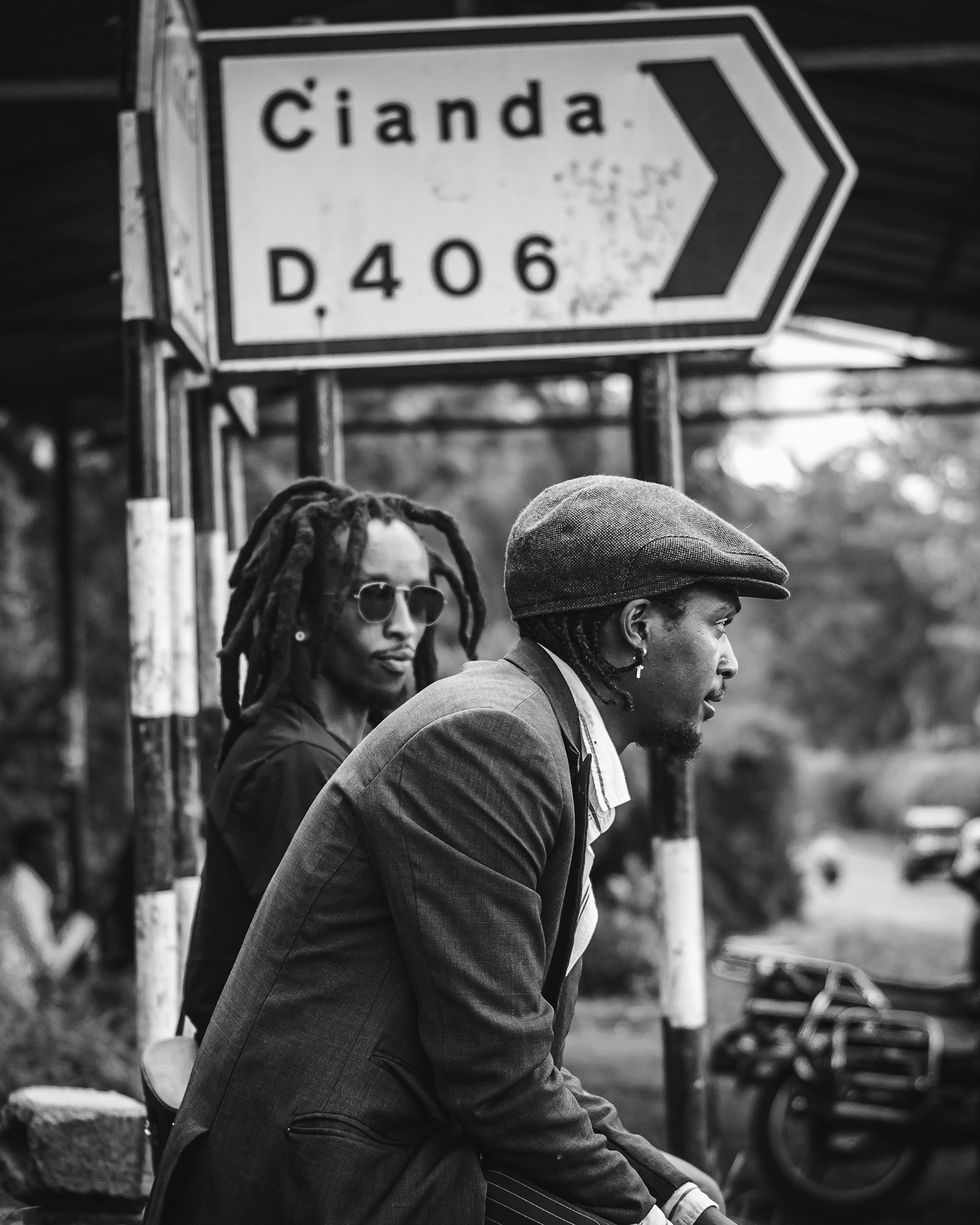 Two men sit by the roadside on Cianda Junction in Kenya. One man with dreadlocks and sunglasses wearing a casual shirt looks at the other man who looks straight ahead and is wearing a sports jacket with a collared shirt and newsboy cap.