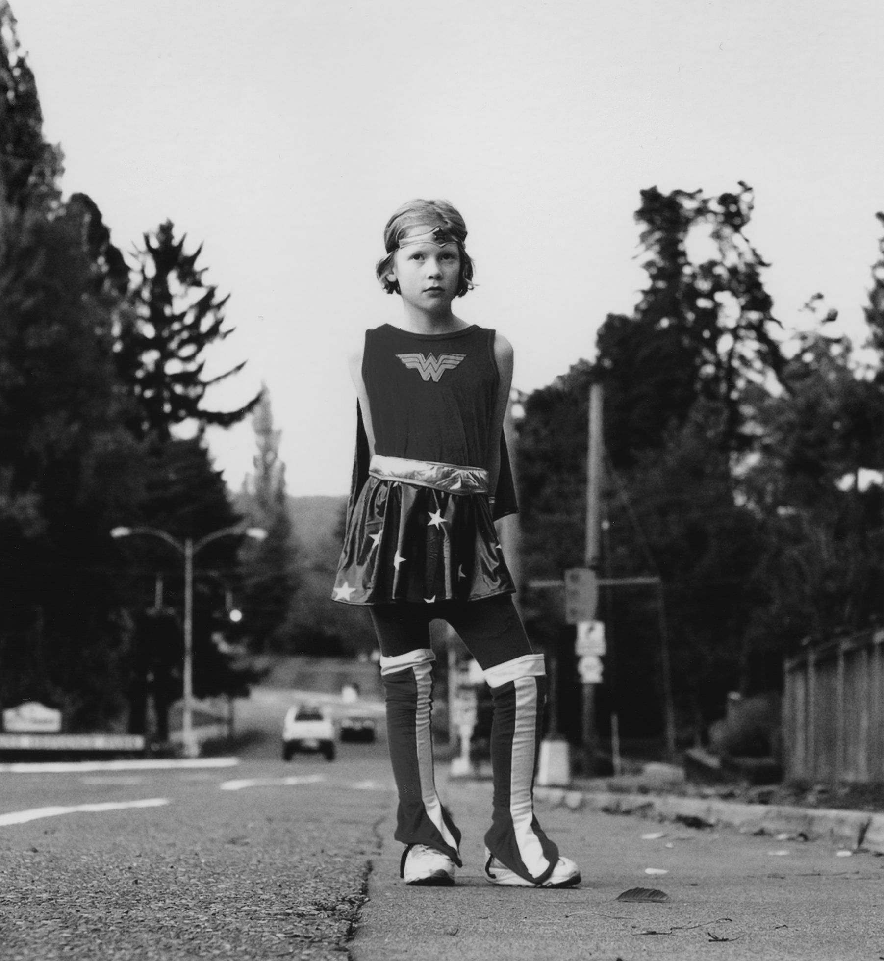 A young girl stands on the side of a road facing the camera with her hands behind her back wearing a Wonder Woman costume complete with cape and headpiece.