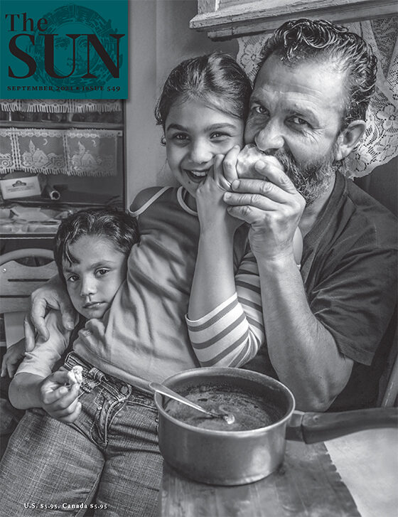 A Romani family. The father is sitting and taking a bite of an apple while his daughter sits on his lap and holds a part of the apple. Another child leans in close to his sister with the father’s other hand seen embracing the second child’s shoulder.