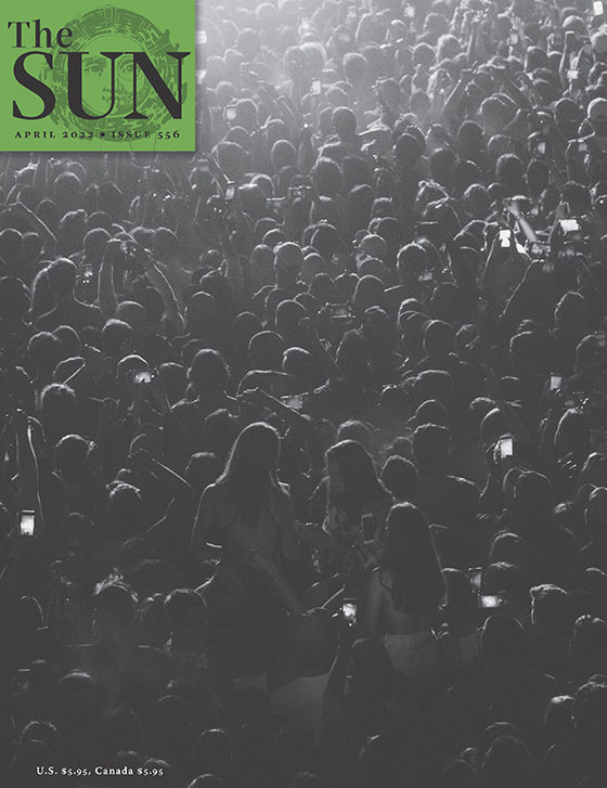 April 2022 cover of The Sun. A 2018 Jack White concert audience in New York City is shown from behind. Concertgoers are standing closely packed in warm-weather gear. The image is dark and the illumination from smartphone screens is sprinkled throughout.