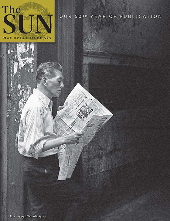 May 2023 cover of The Sun. A man stands in front of the entrance to his building in Manhattan’s Chinatown in 1975 while reading a newspaper. The man is wearing dress pants and a button-down shirt with the sleeves rolled up to his elbows.